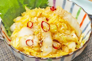 白菜と唐辛子の漬物がボウルに盛られている。ゴマがトッピングされており、バックに葉が添えられている。