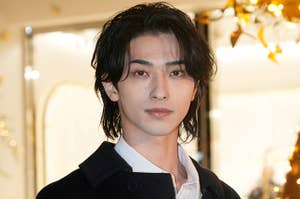 男性がイベントでシンプルなコートとシャツを着てポーズしています。背後には光がきらめいています。