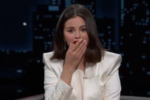 A woman with shoulder-length hair in a white blazer covers her mouth with her hand, appearing surprised, against a cityscape background