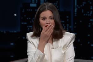 A woman with shoulder-length hair in a white blazer covers her mouth with her hand, appearing surprised, against a cityscape background