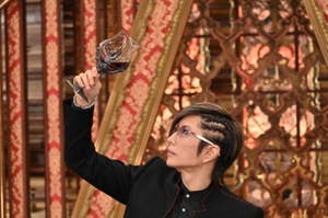 男性がゴージャスな背景を背にワイングラスを持ち上げている。黒のジャケットを着用。
