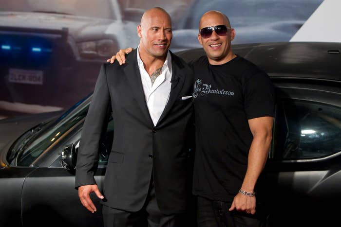 Two celebrities pose together; one is in a sleek suit, the other in a casual t-shirt and sunglasses, standing in front of a car backdrop