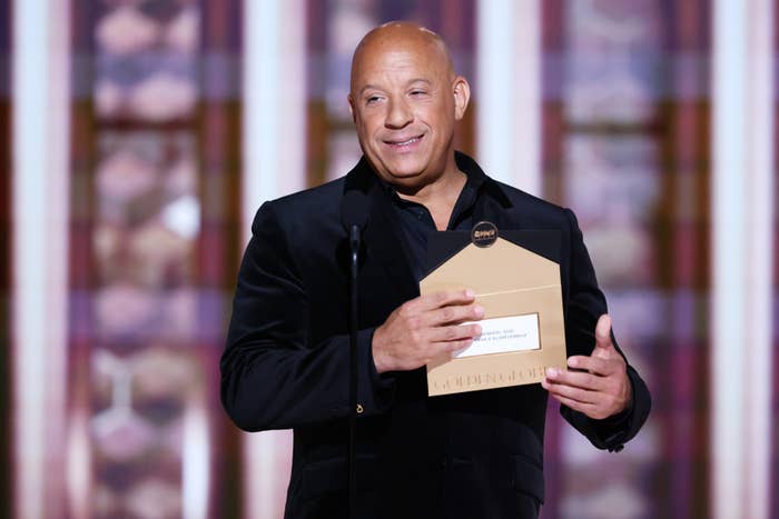 Man in a velvet suit onstage holds a paper, smiling, possibly announcing an award at a celebrity event