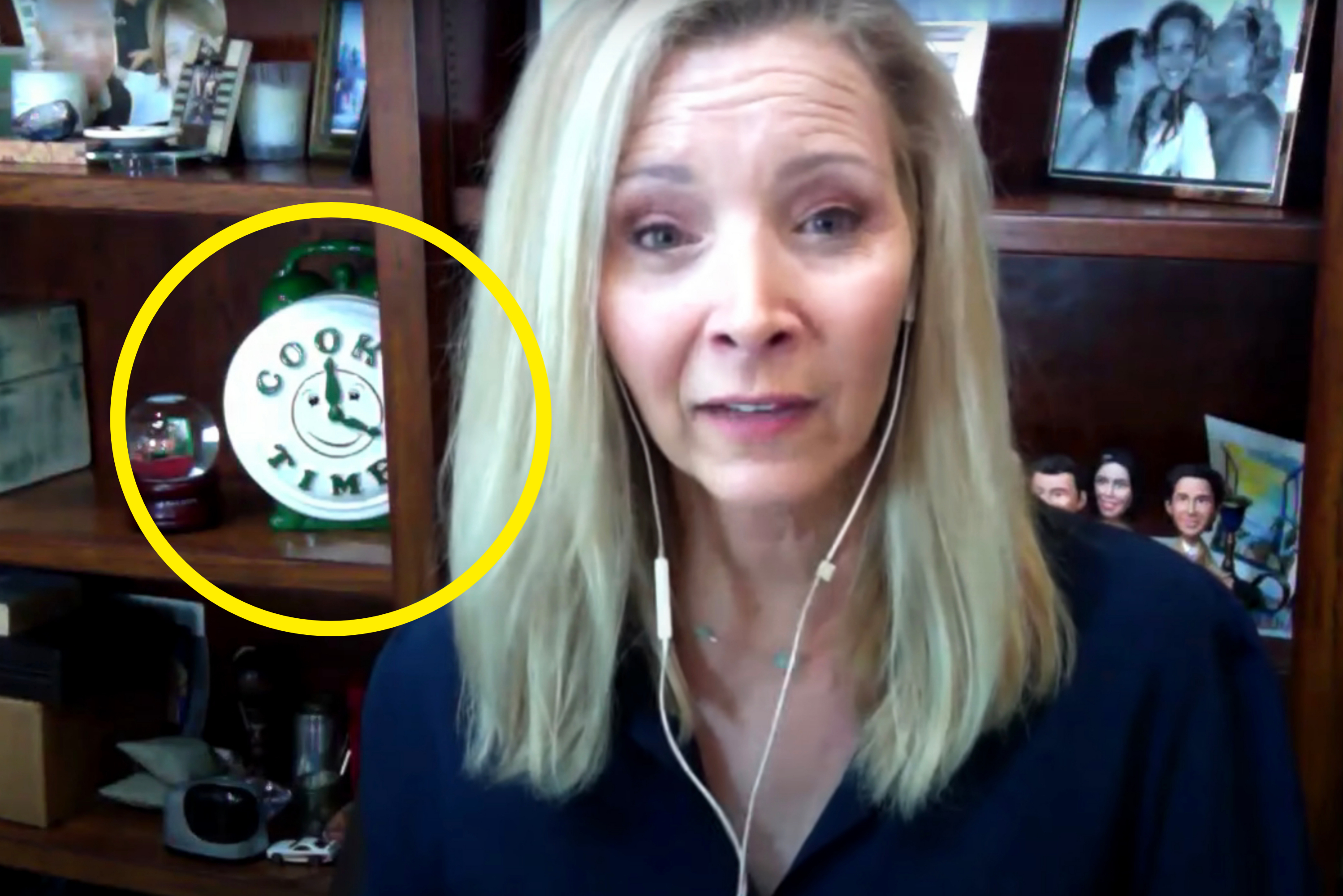Person with headphones, seated in front of a bookshelf with photos and a clock, speaking in a video or virtual meeting setting