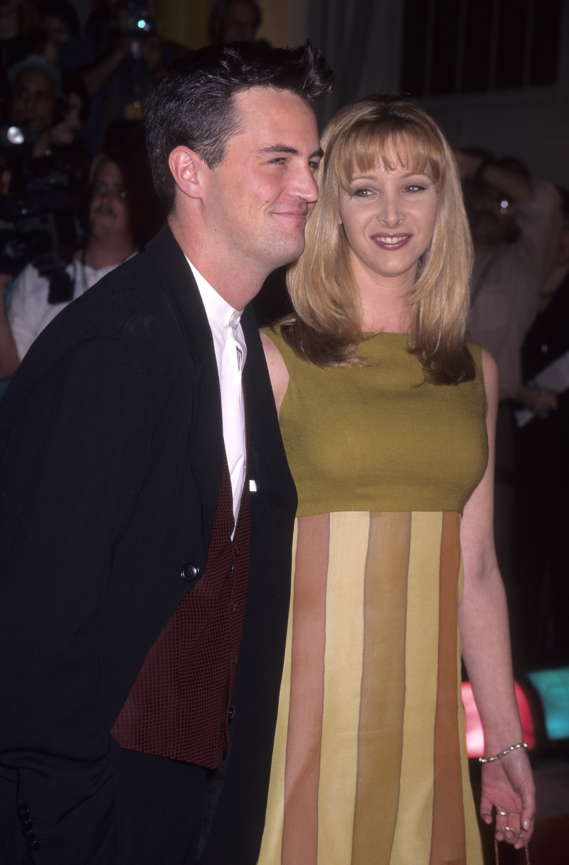 Two celebrities on a red carpet; man in a suit with a vest, woman in a sleeveless striped dress, smiling for photographs