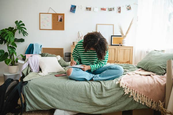 Person sitting cross-legged on a bed, writing in a notebook, surrounded by cozy decor and plants