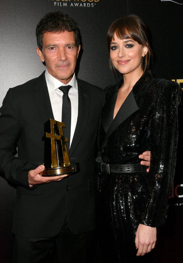 Two people dressed in formal black clothing pose for a photo. One holds a film award trophy