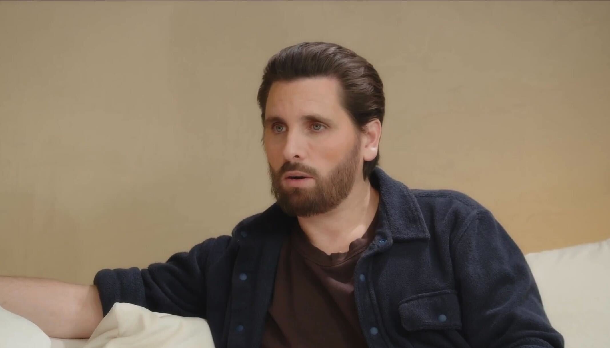 Man seated, wearing a casual dark jacket and t-shirt on a cream couch, appearing engaged in conversation
