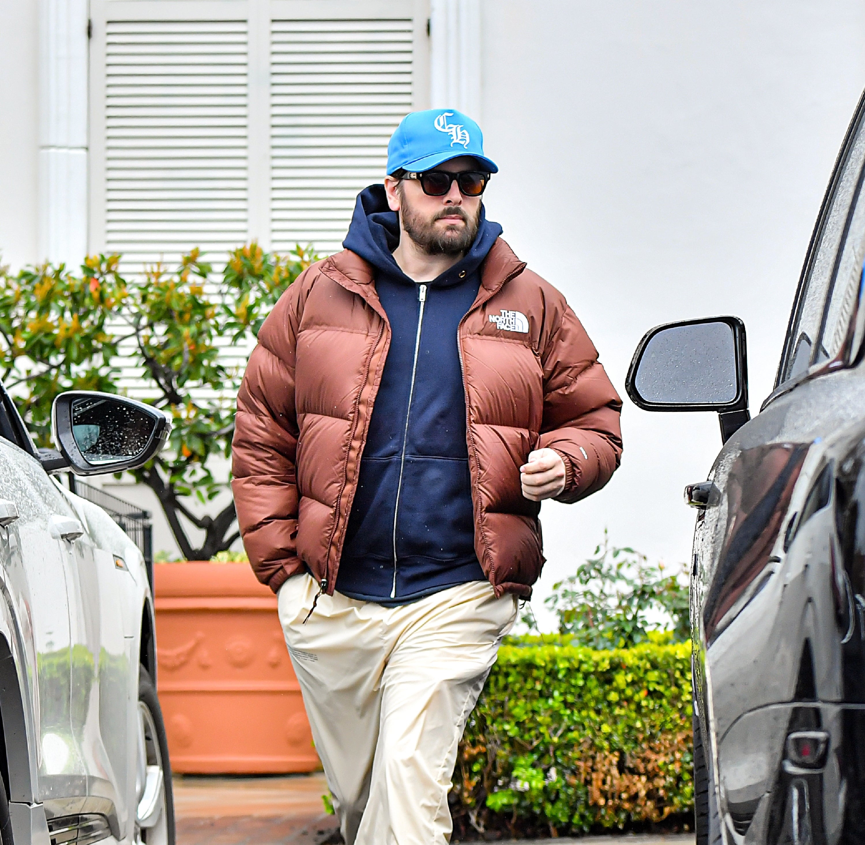 Person wearing a puffer jacket, hoodie, sunglasses, and a cap walks between cars. Casual celebrity street style