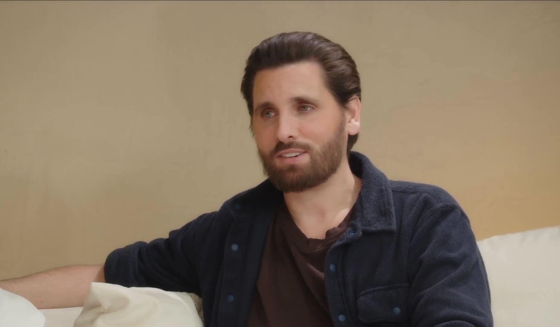 A person with neatly styled hair and a beard, wearing a dark casual jacket, sits on a couch, engaged in conversation