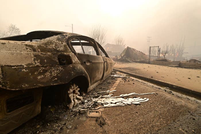 A burnt-out car on a road amidst destruction, with charred debris and a hazy sky in the background, indicating aftermath of a fire