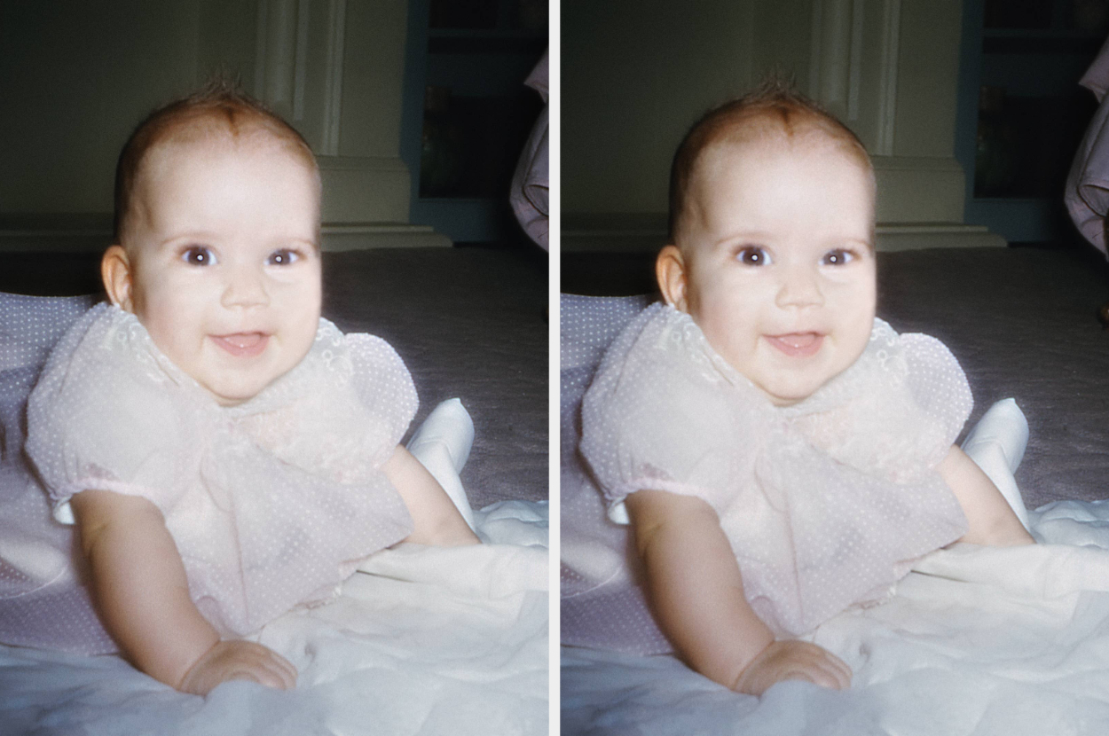 A smiling baby in a light dress lies on a soft surface indoors, facing forward