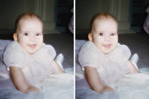 A smiling baby in a light dress lies on a soft surface indoors, facing forward