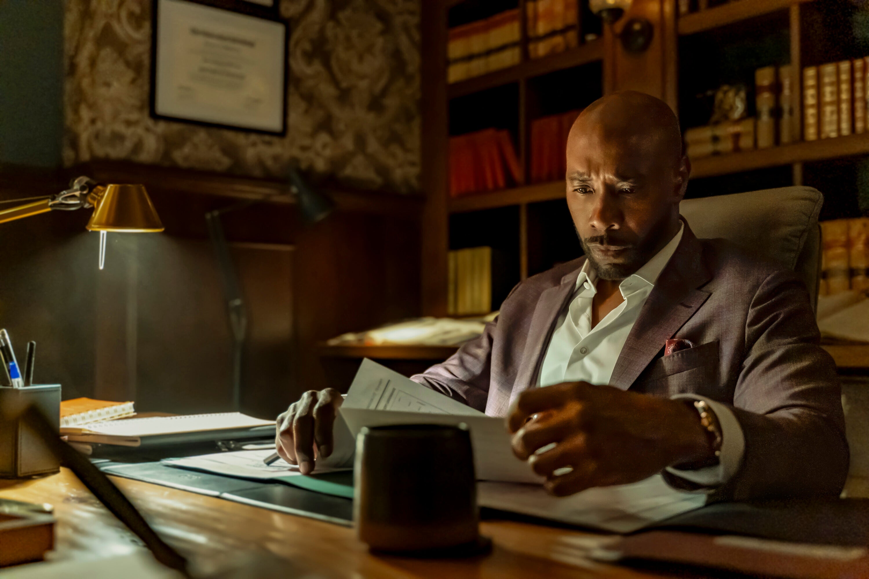 A man in a suit is sitting at a desk, focused on reading documents, with shelves of books in the background