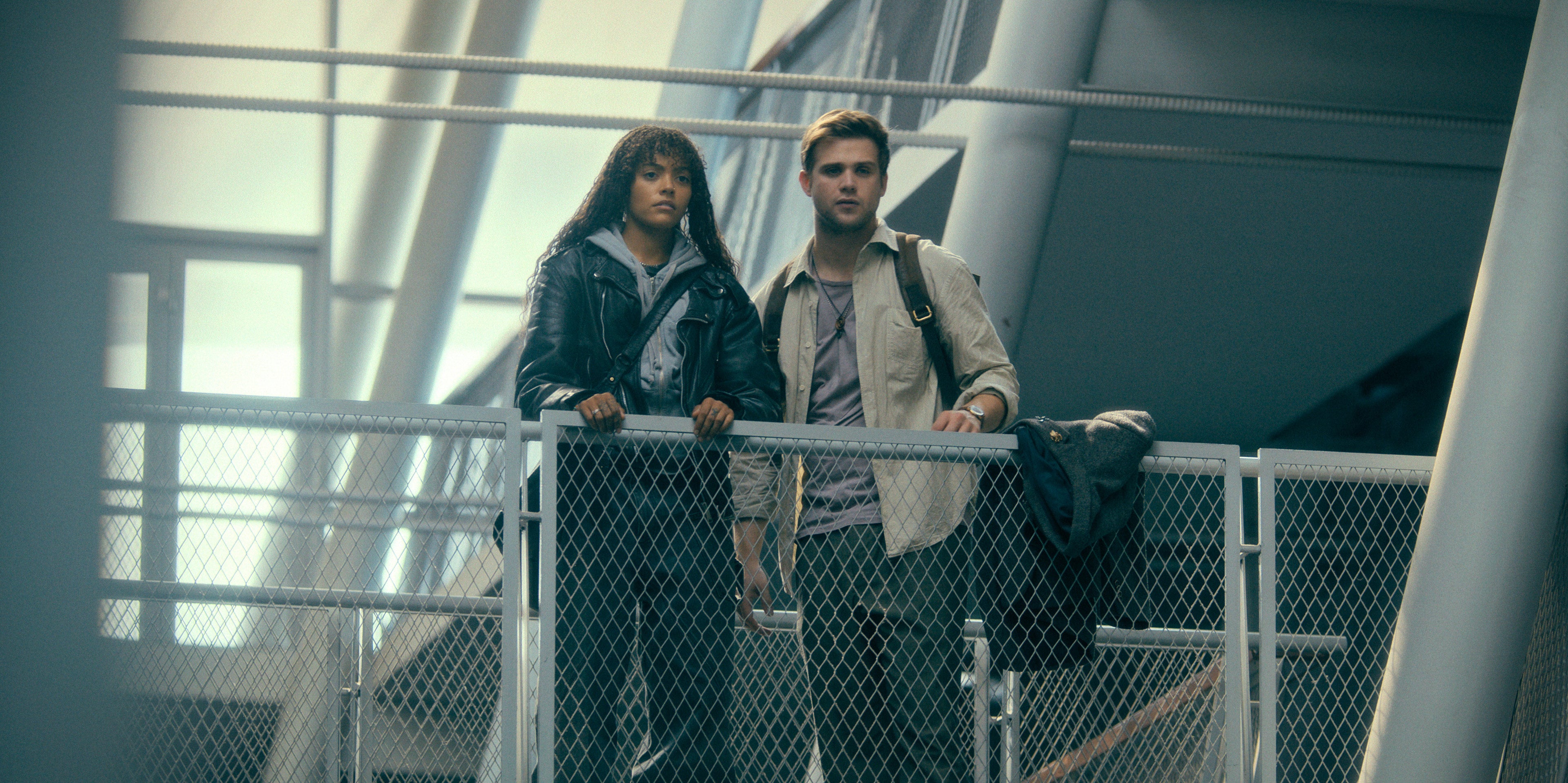 Two people stand on a metal bridge with serious expressions, dressed in casual, modern attire