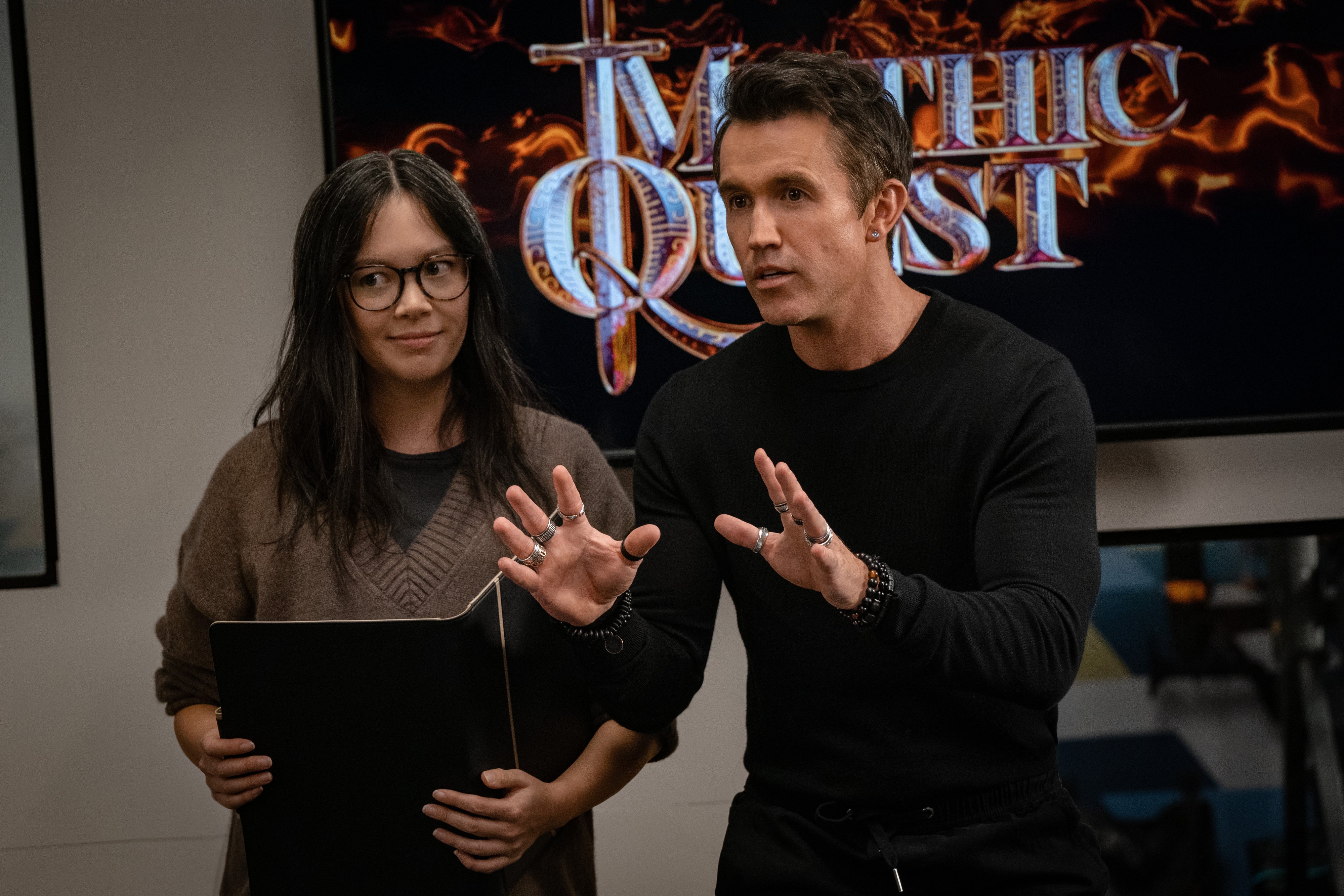 Two people are discussing in front of a "Mythic Quest" sign. One holds a folder, and the other is gesturing with their hands