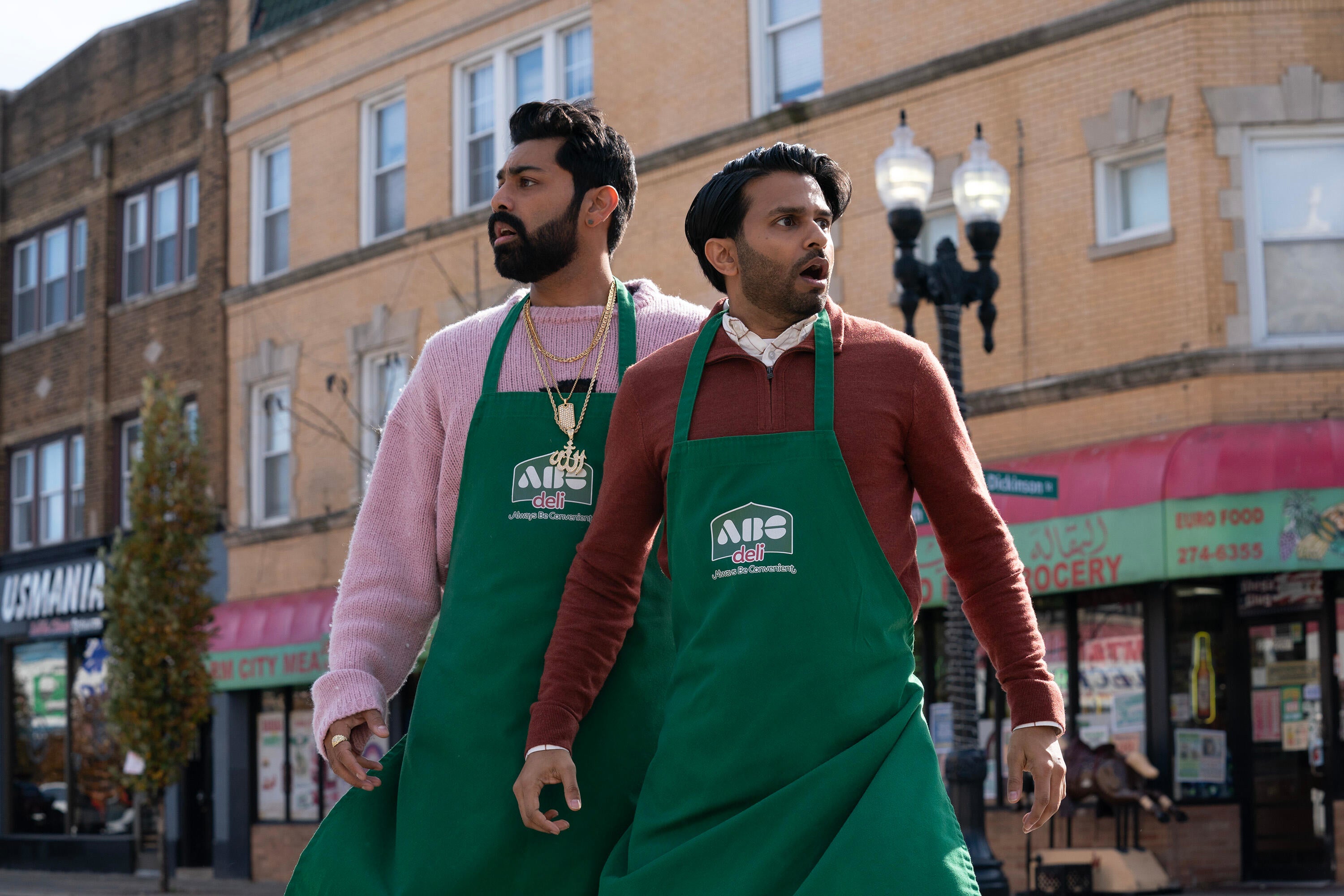 Two actors in green aprons stand on a street, looking surprised, a scene from a TV show or movie