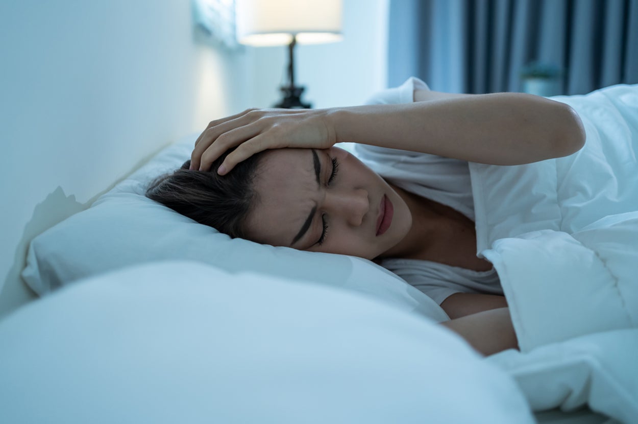 Person lying in bed, holding their head in discomfort, suggesting a headache or stress. Bedroom setting with a bedside lamp in the background