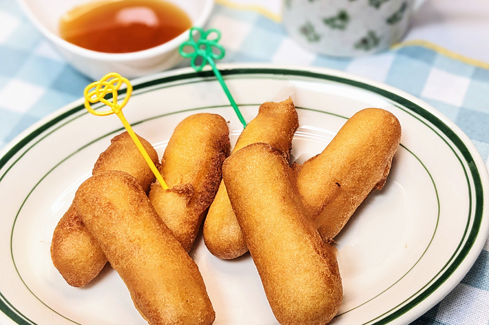 皿に盛られた揚げドッグとハチミツが置かれたテーブル。背景に花柄のカップ。