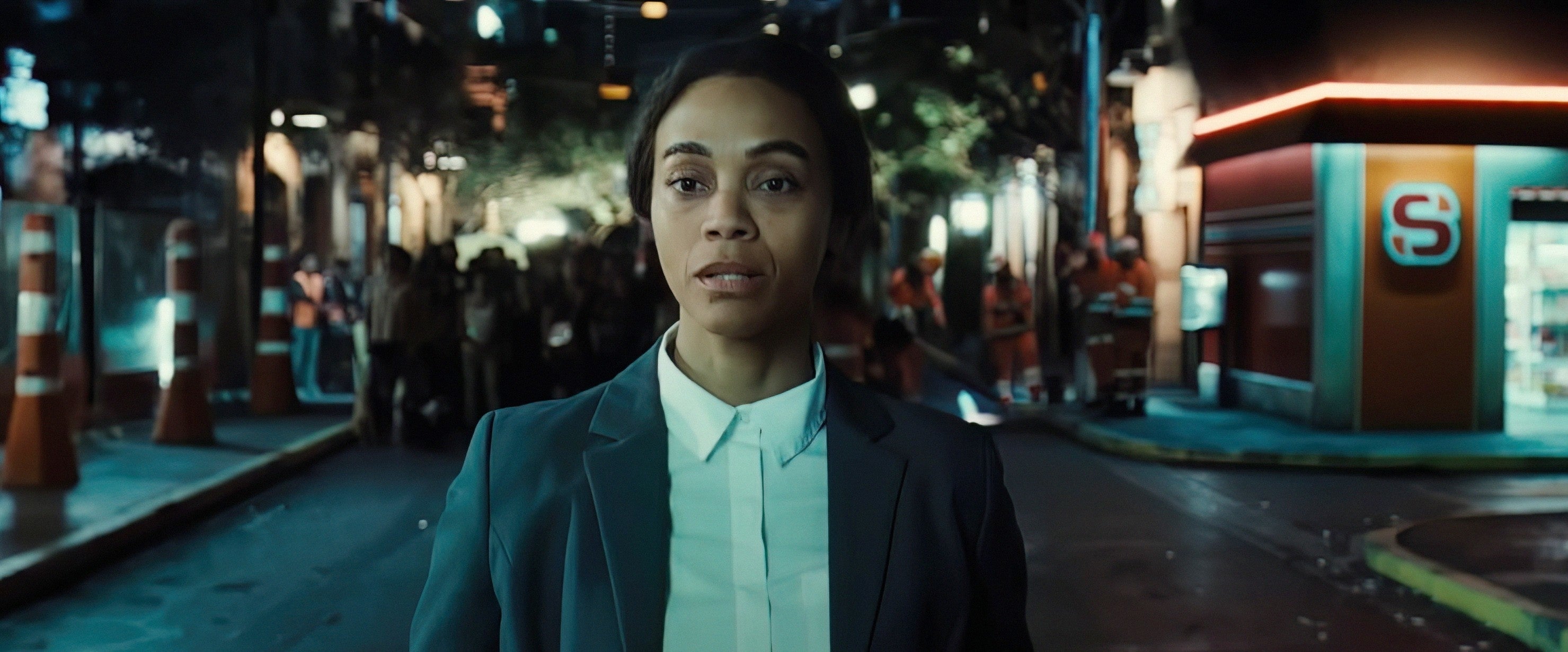 Zoe Saldana in a business suit walks down a city street at night, with a serious expression, against a backdrop of blurred buildings and streetlights