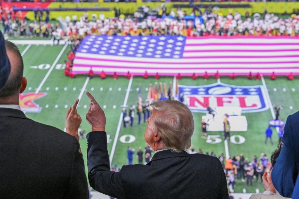 A person in a suit gestures from a stadium box over an NFL field with a large American flag displayed and teams lined up