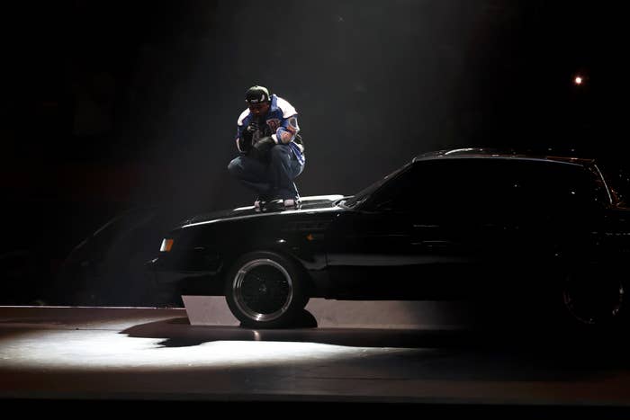 Kendrick Lamar squatting on a car during a dramatic stage performance, wearing casual streetwear and a cap