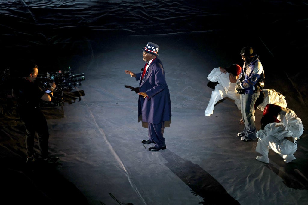 A person in a stylish long coat and a star-patterned hat performs on stage with dancers and a camera operator nearby