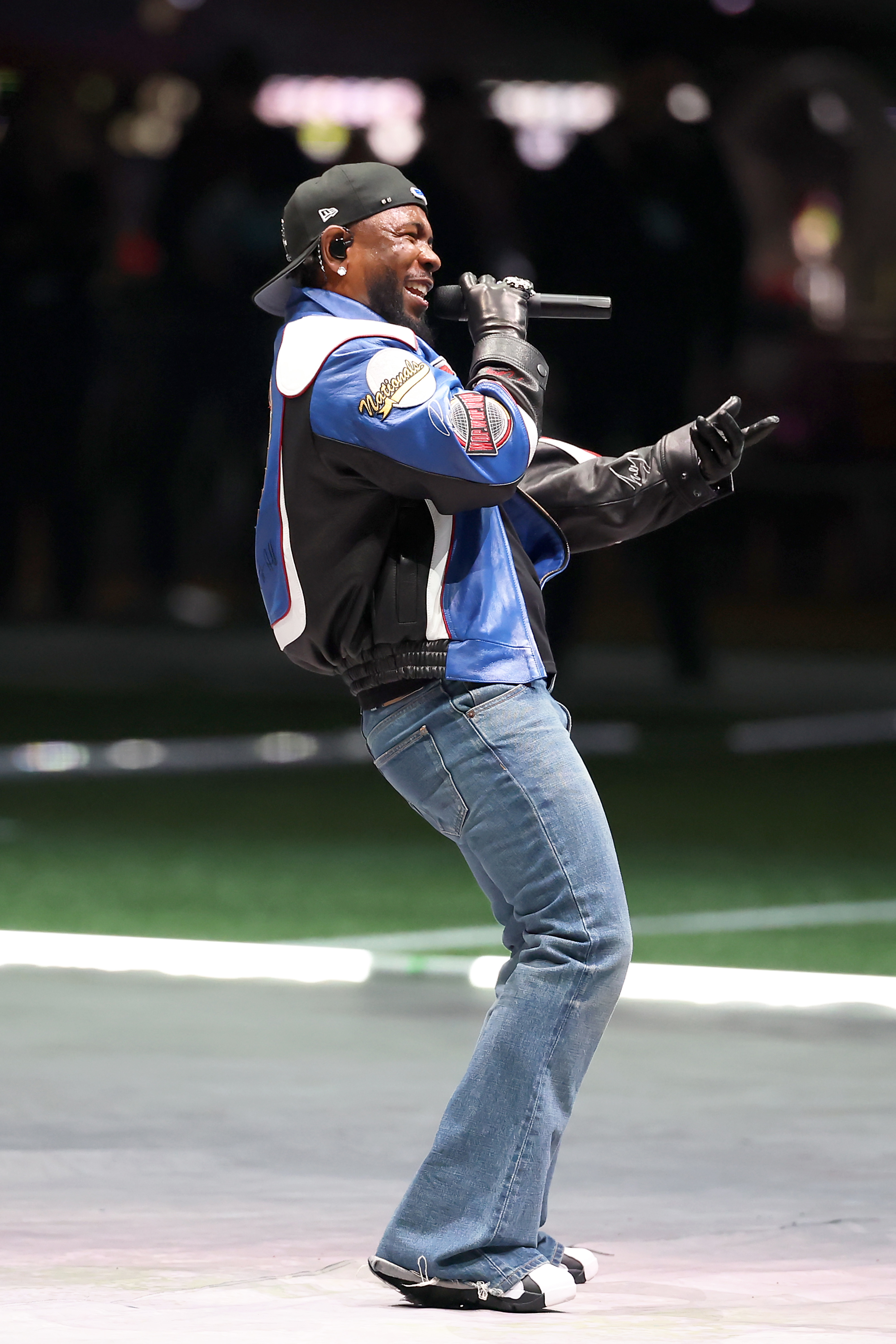 Kendrick Lamar wearing jeans, letterman jacket, cap, and gloves, singing on stage with a microphone