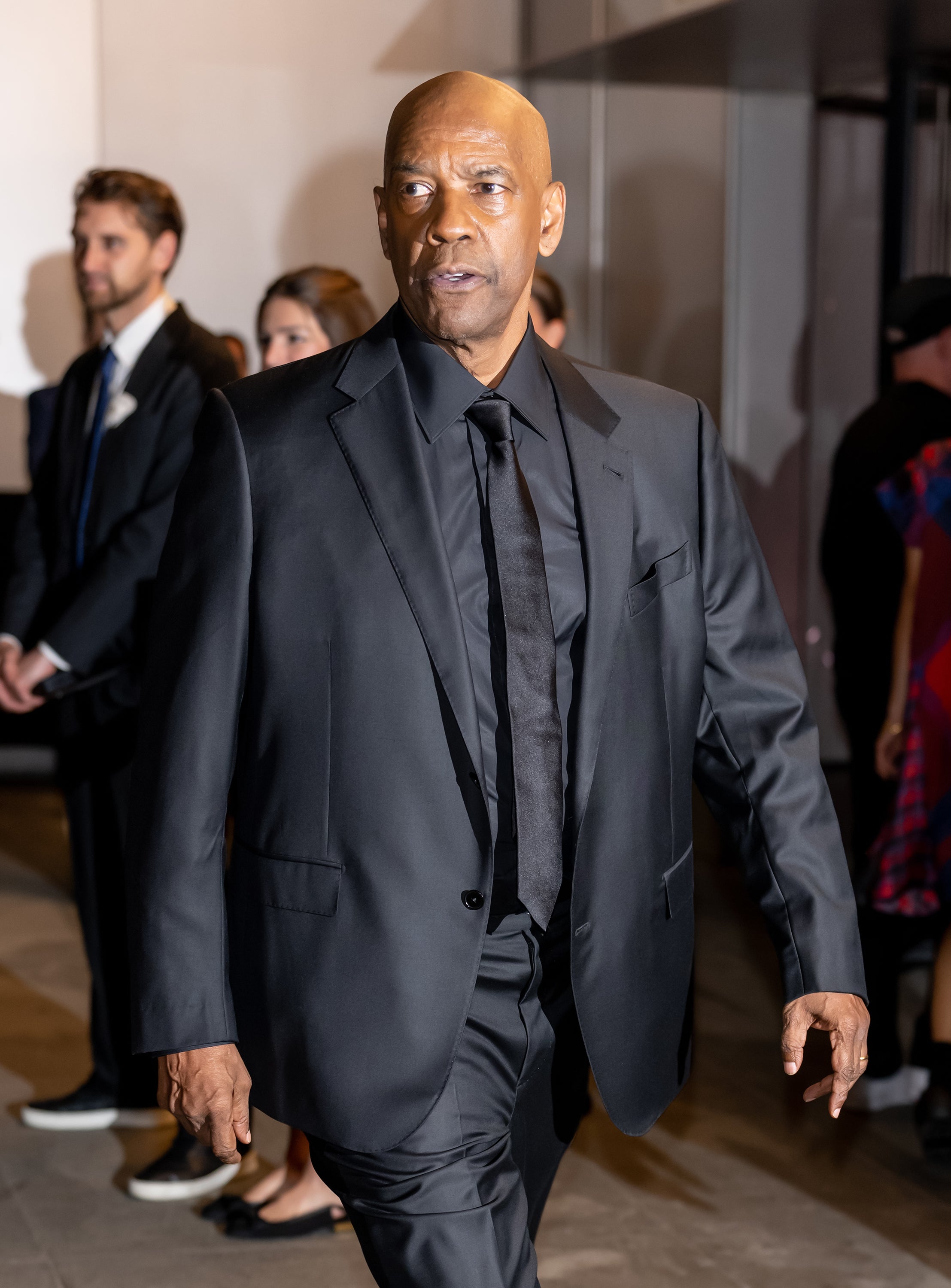Denzel Washington in a sleek black suit walking at an event, surrounded by other attendees