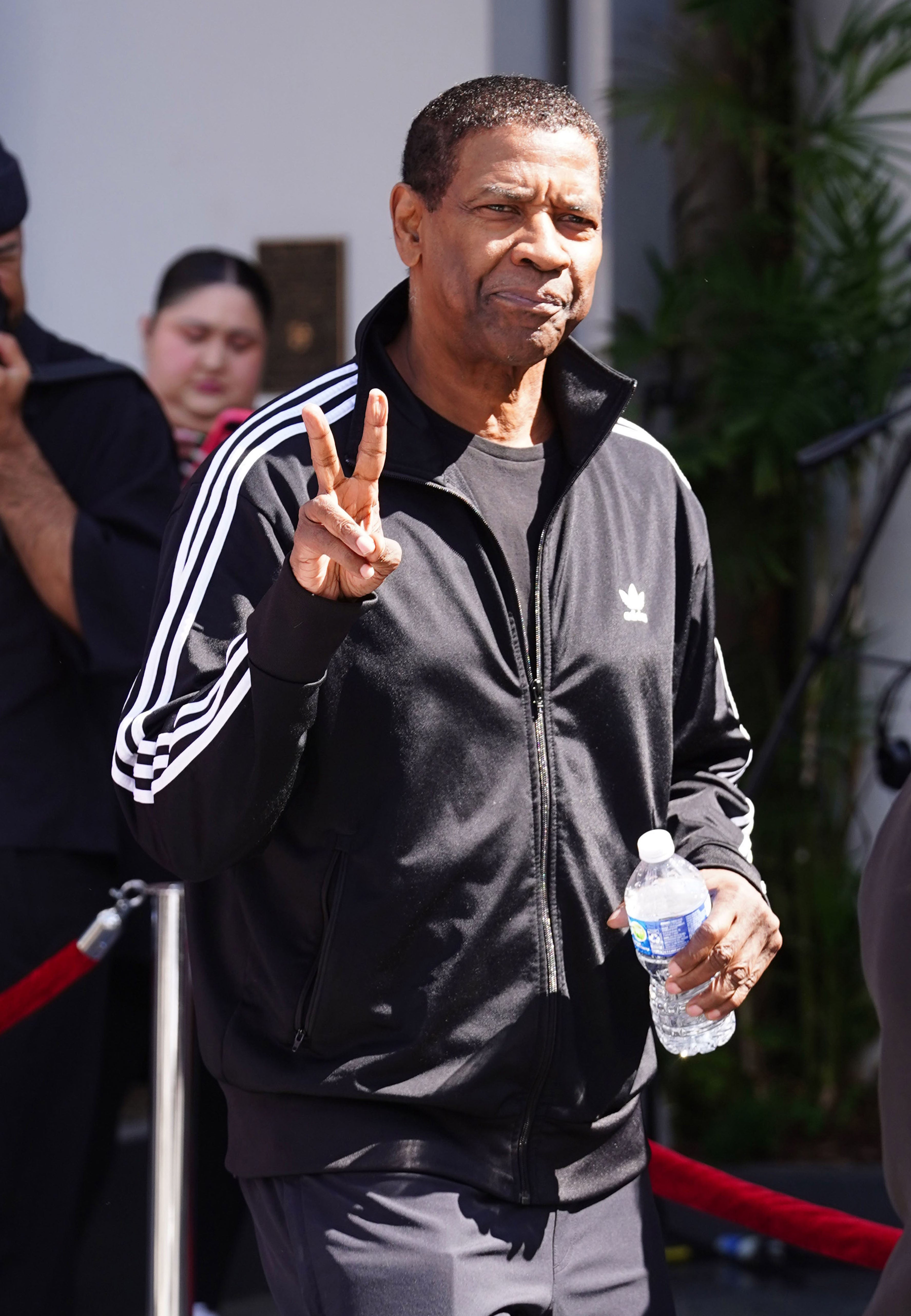 Denzel Washington in an Adidas tracksuit holds up a peace sign while holding a water bottle