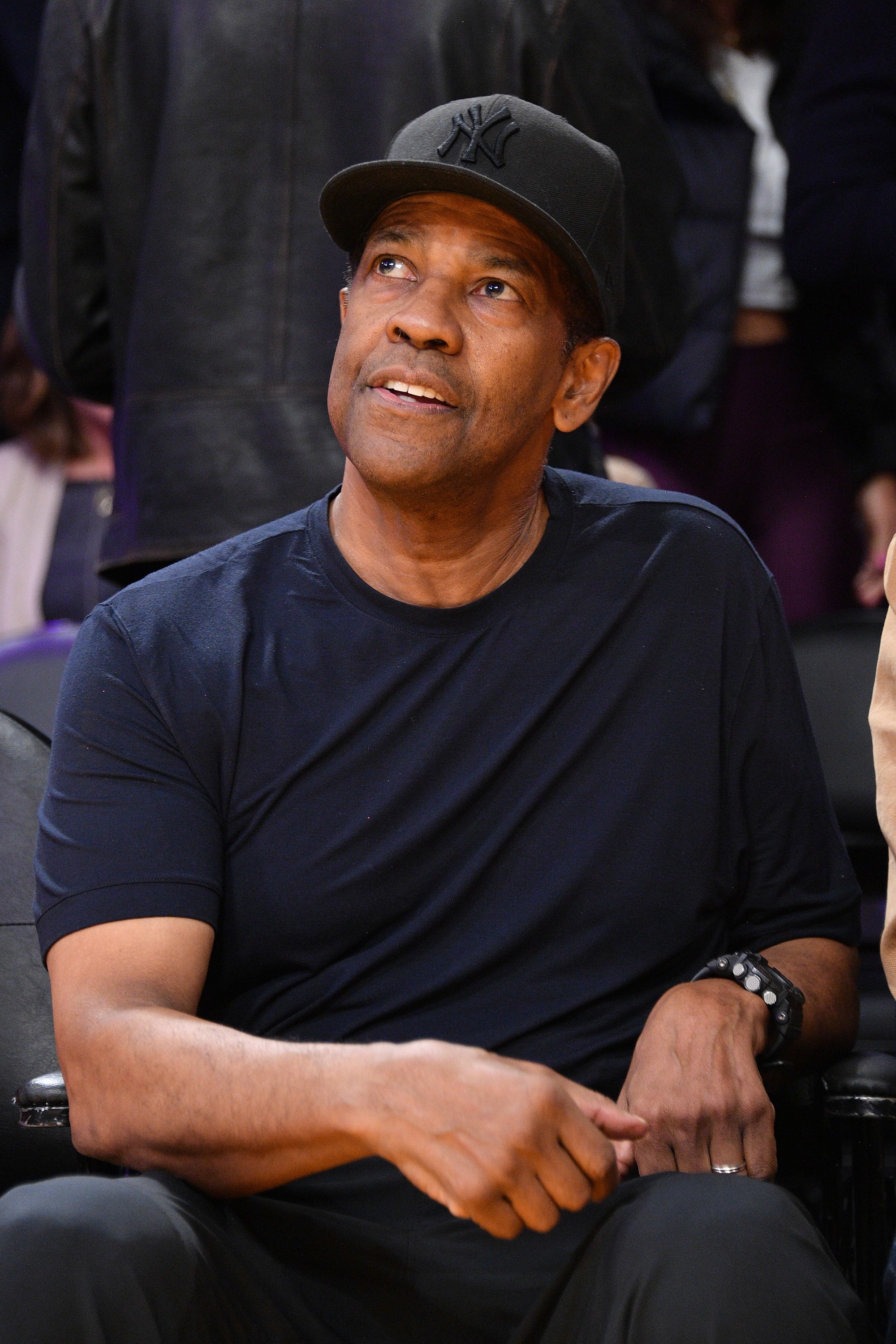 Denzel Washington seated courtside at a basketball game, wearing a casual black cap and shirt, looking upwards