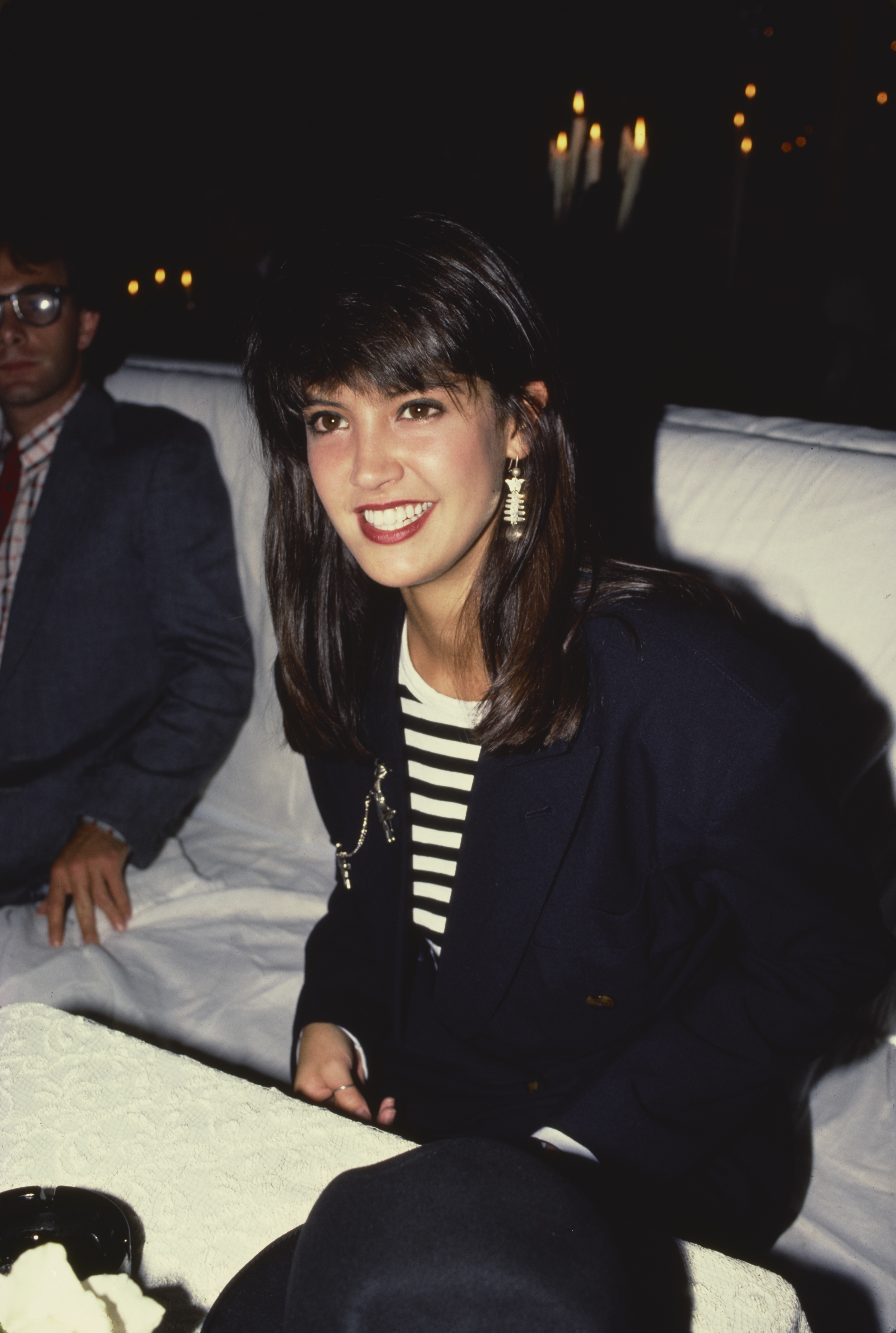 Person smiling at an event, dressed in a striped shirt with a dark blazer, seated indoors. Candles are visible in the background