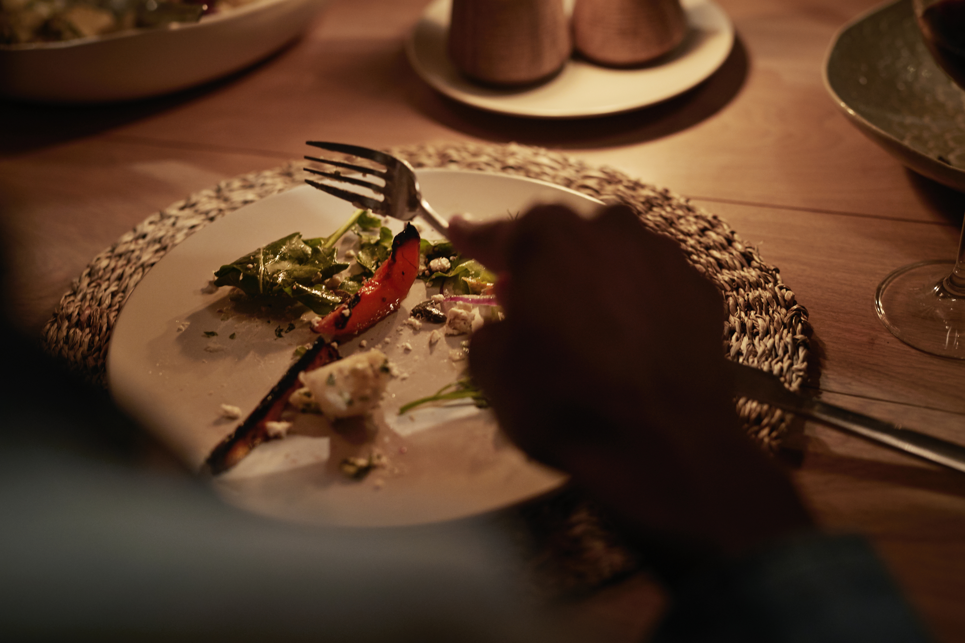 Person spiser, holder en gaffel over en tallerken med delvis spist salat og grillede grønnsaker på en vevd placemat, og skaper en koselig, intim atmosfære