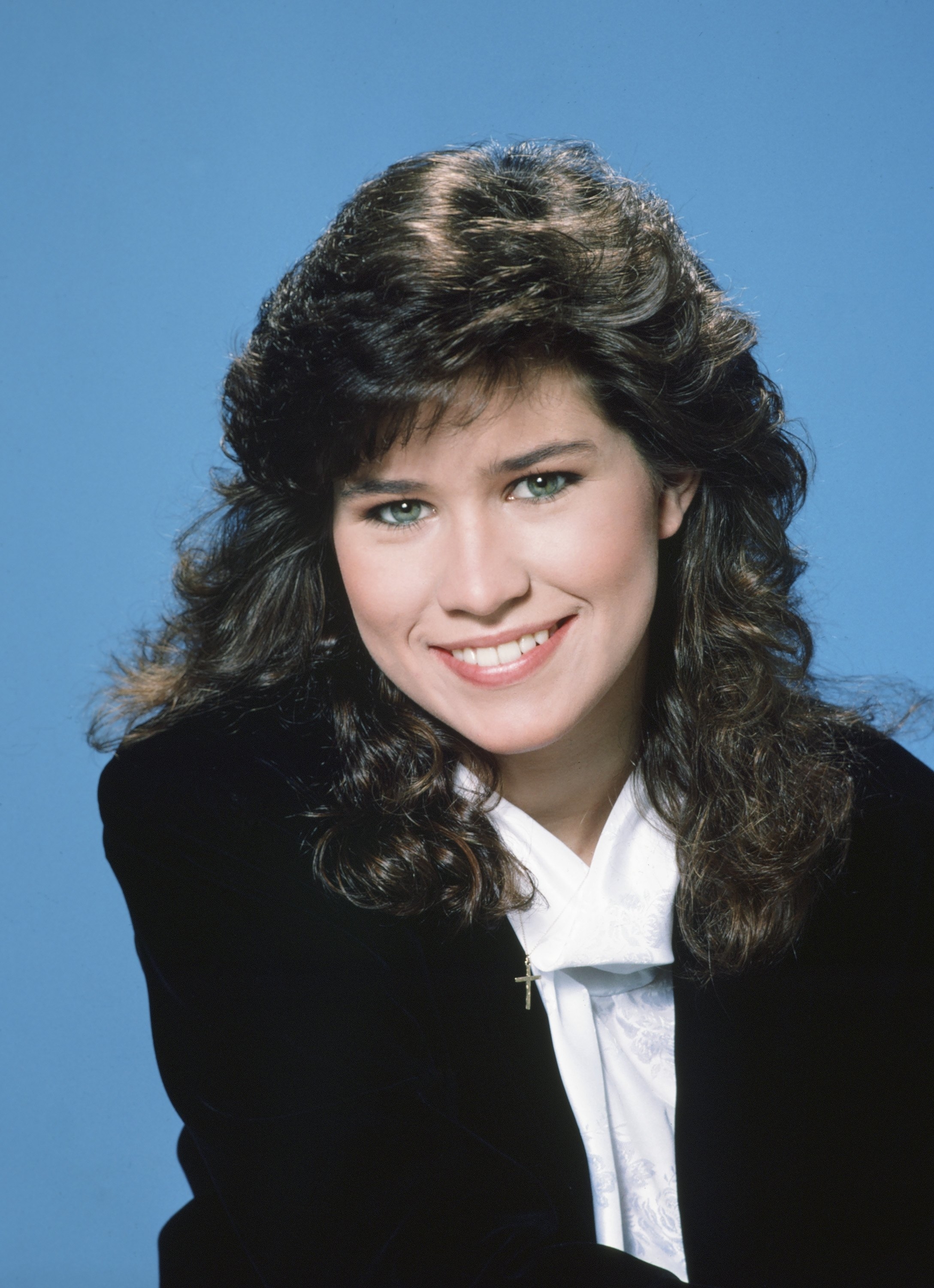 Person with long, wavy hair smiles, wearing a black jacket over a white shirt
