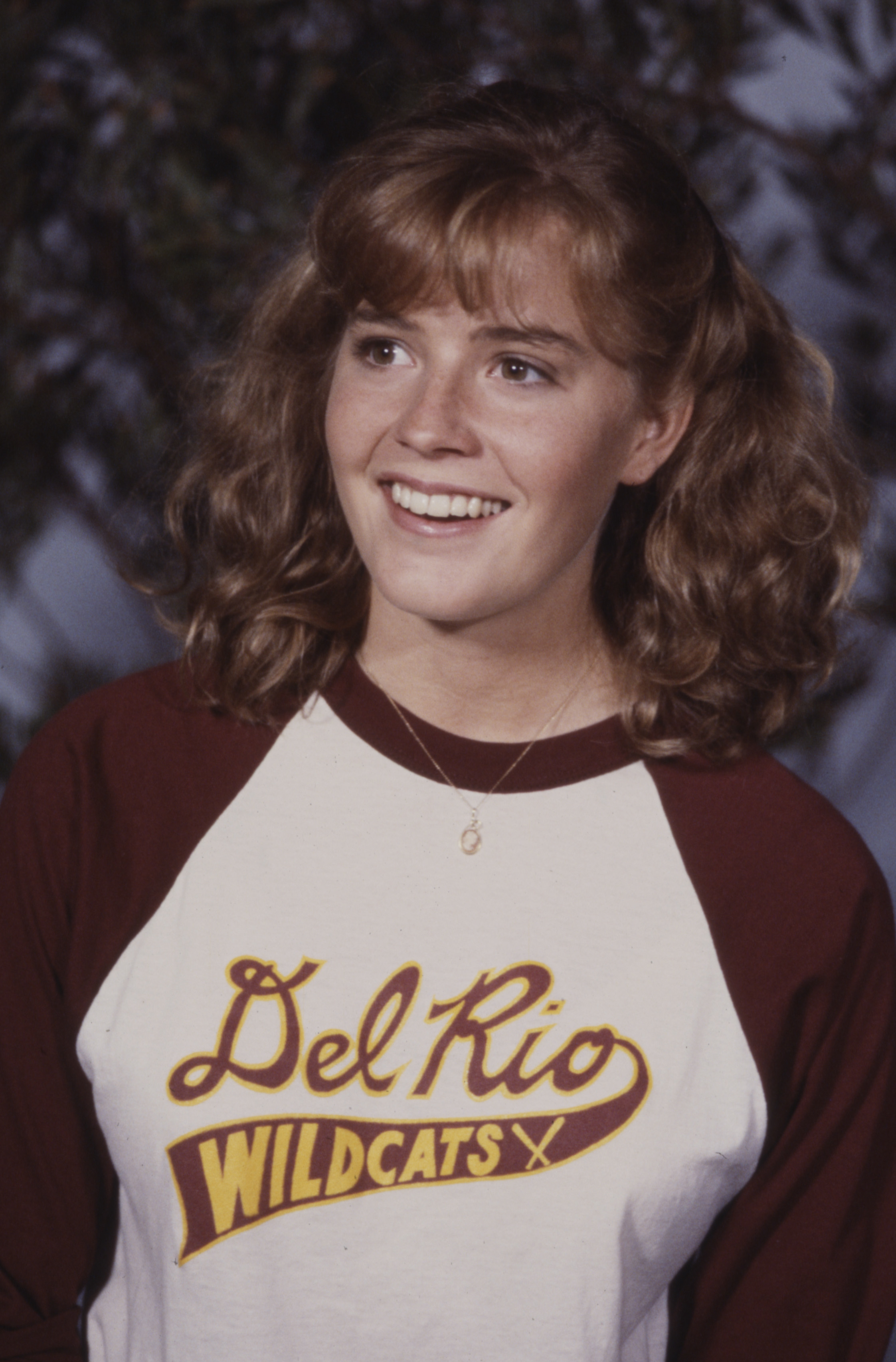 Person smiling in a &quot;Del Rio Wildcats&quot; jersey with curly hair styled in pigtails, against a blurred background