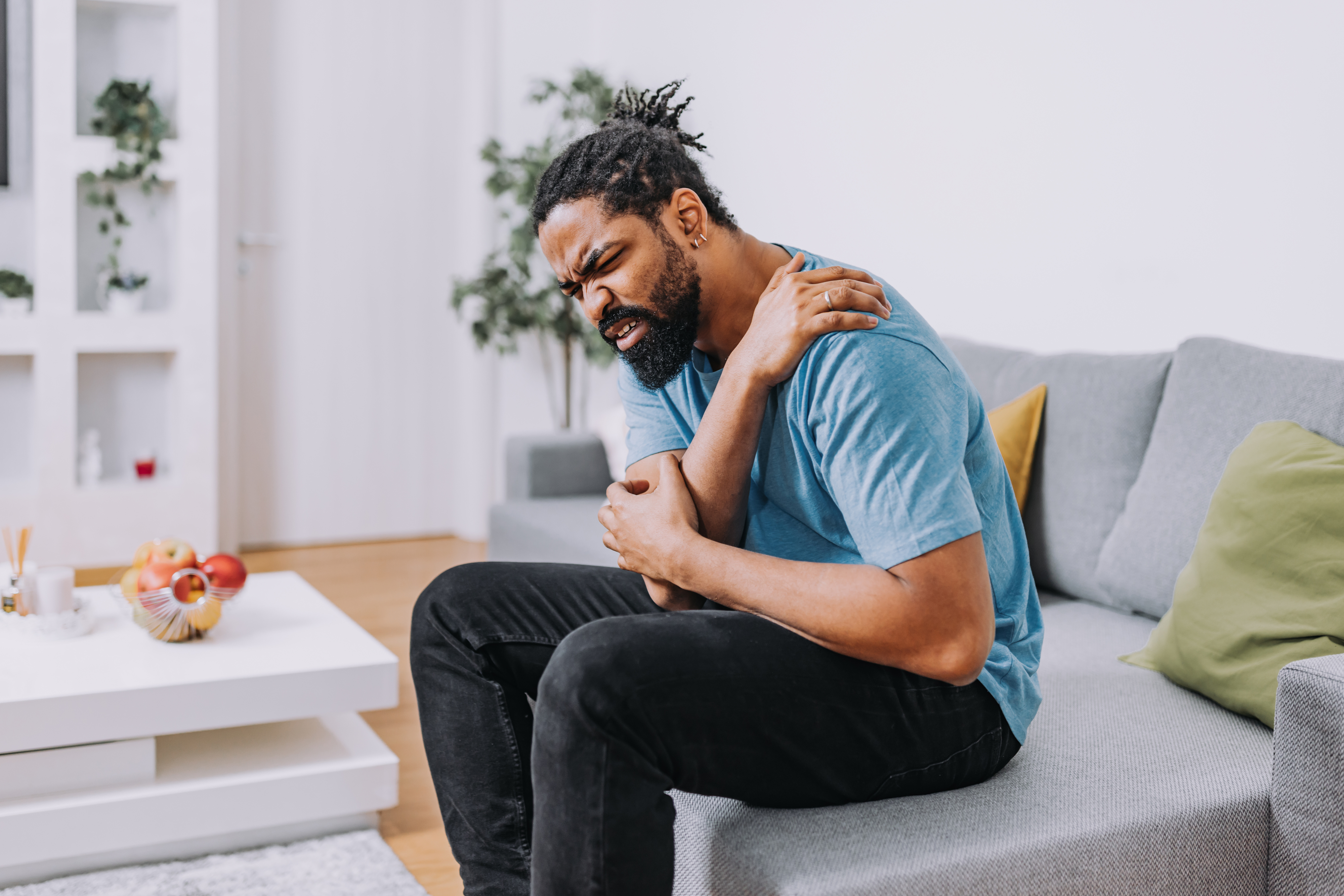 Mannen som sitter på en sofa holder skulderen og vinner i smerter, har på seg en uformell blå skjorte og svarte bukser. En skål med frukt er på bordet i nærheten