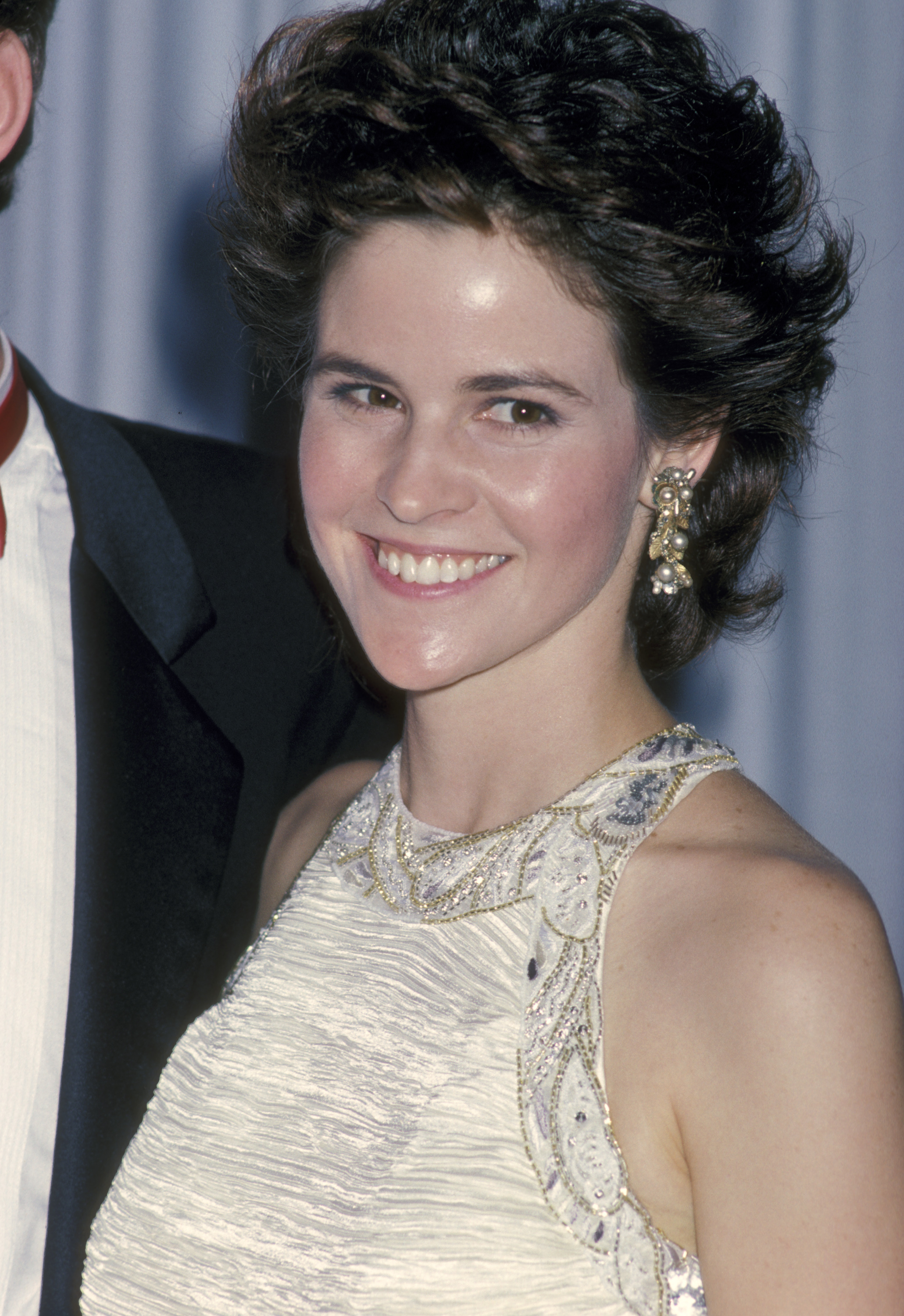 A woman in a sleeveless, bejeweled gown and large earrings smiles at a formal event