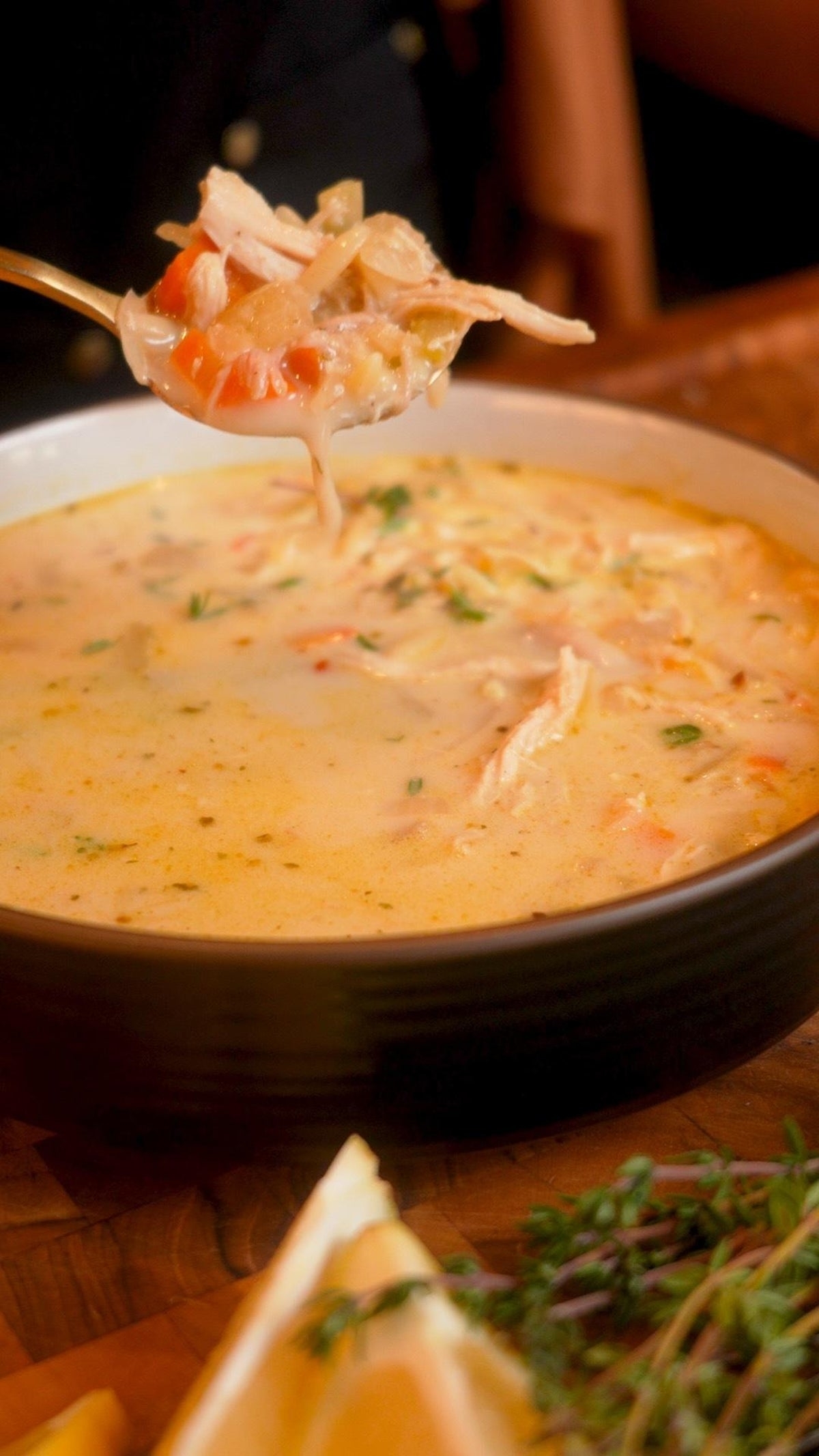 A spoonful of creamy soup with chunks of chicken and vegetables is held above a bowl, with herbs and lemon wedges nearby