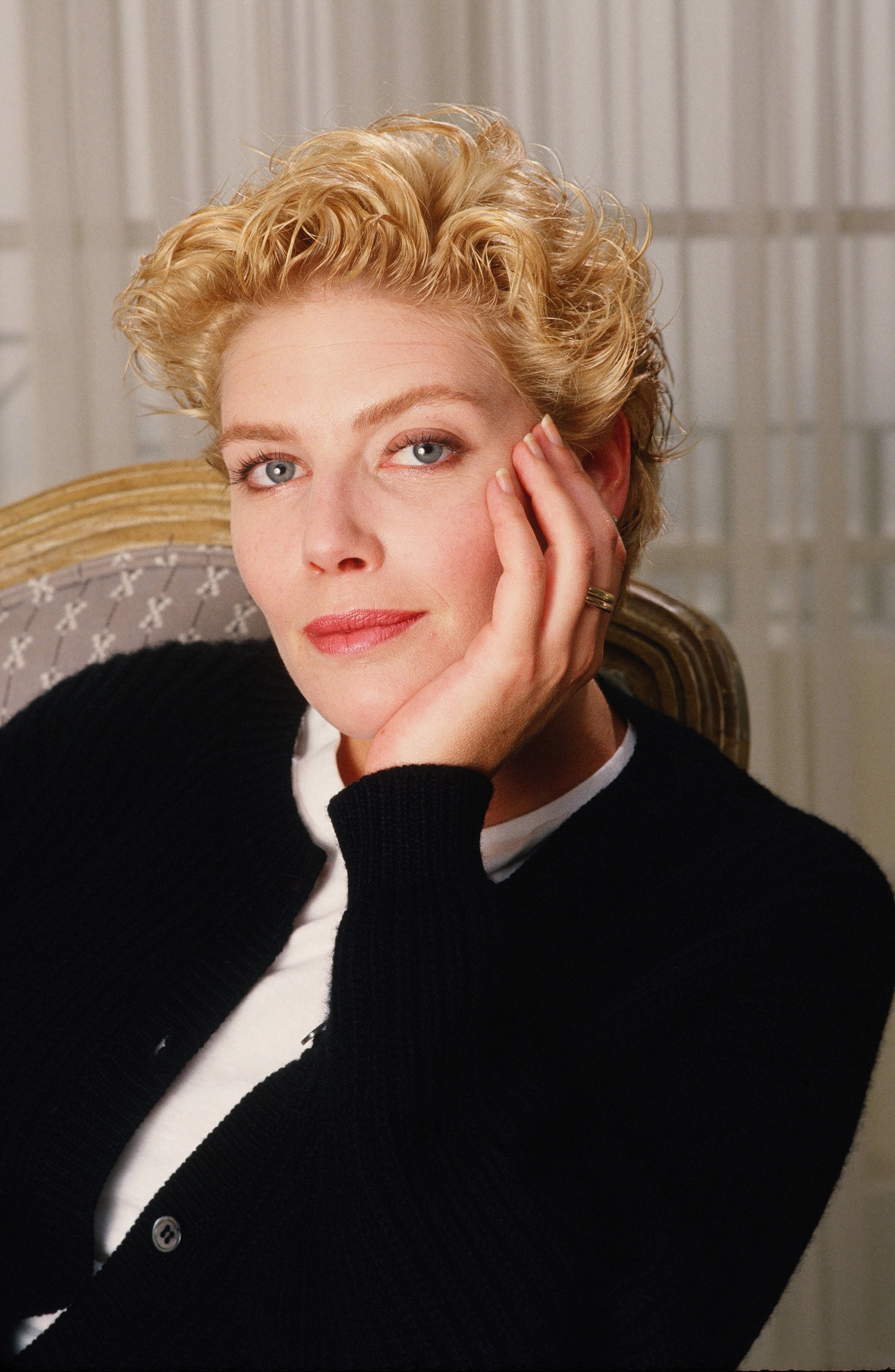A person with short, styled hair sits in a chair, wearing a black cardigan over a white top, resting their head on their hand and looking at the camera