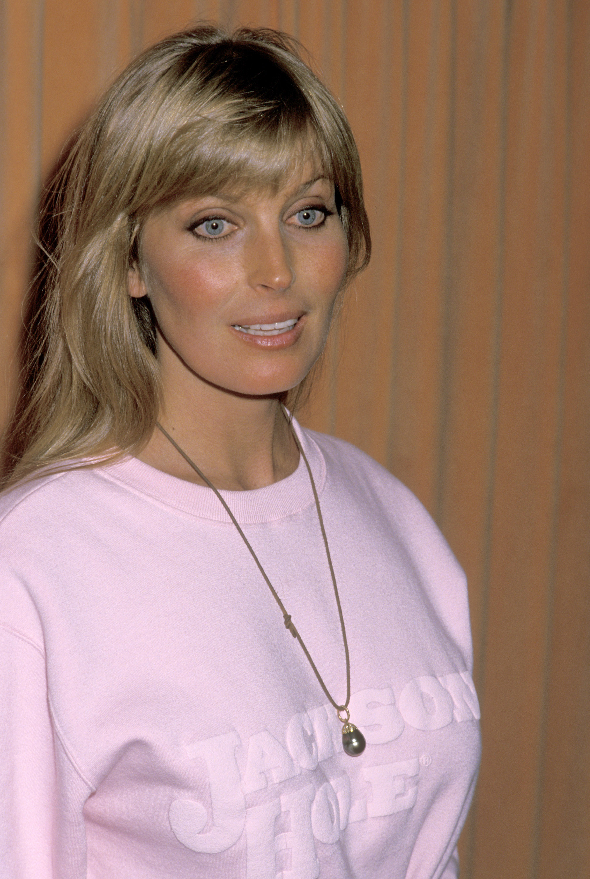 Person with long hair wearing a necklace and a sweatshirt with &quot;Jackson Hole&quot; text, standing in front of a plain background