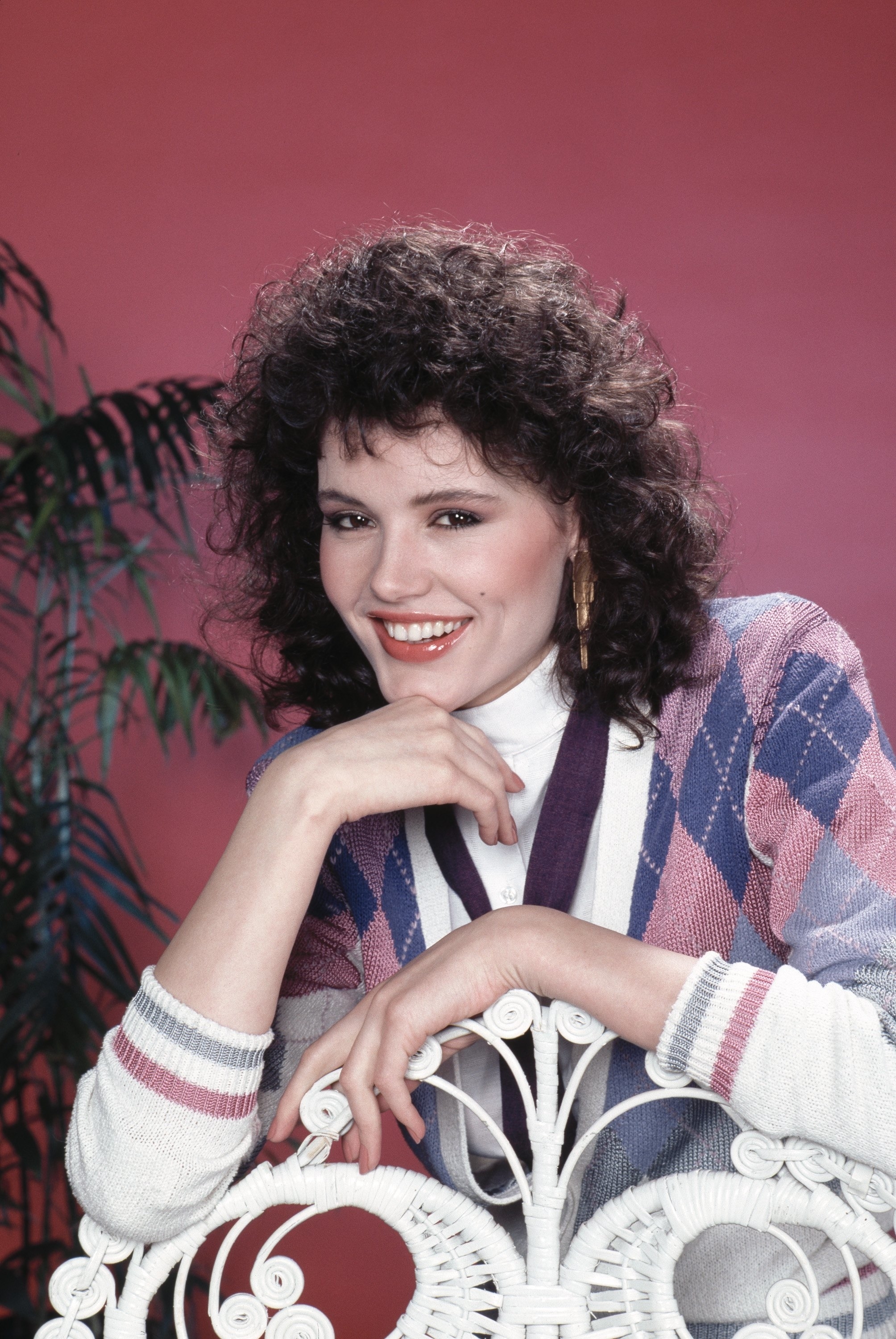 Person with curly hair smiles, wearing an argyle-patterned sweater over a white turtleneck, posing against a decorative chair in a studio setting