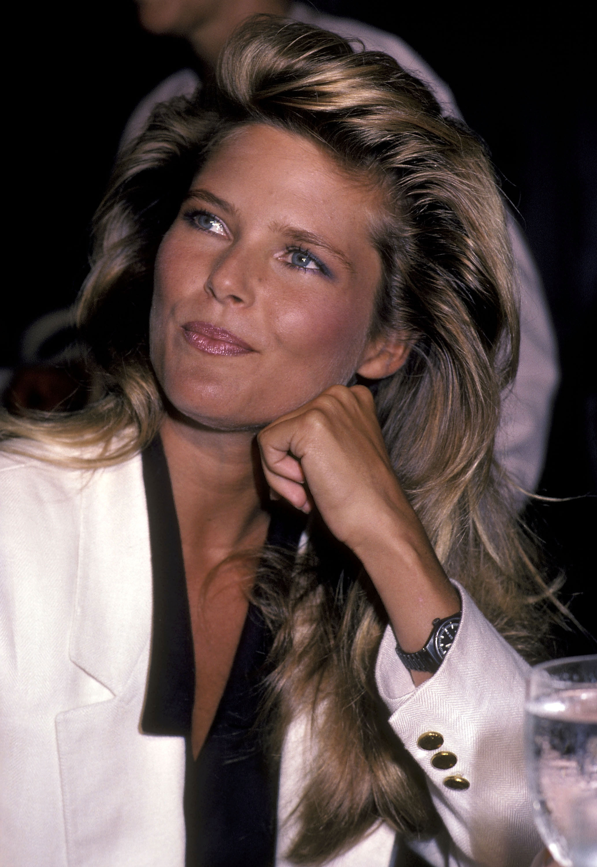 Person in fashionable blazer with long hair, resting chin on hand at event