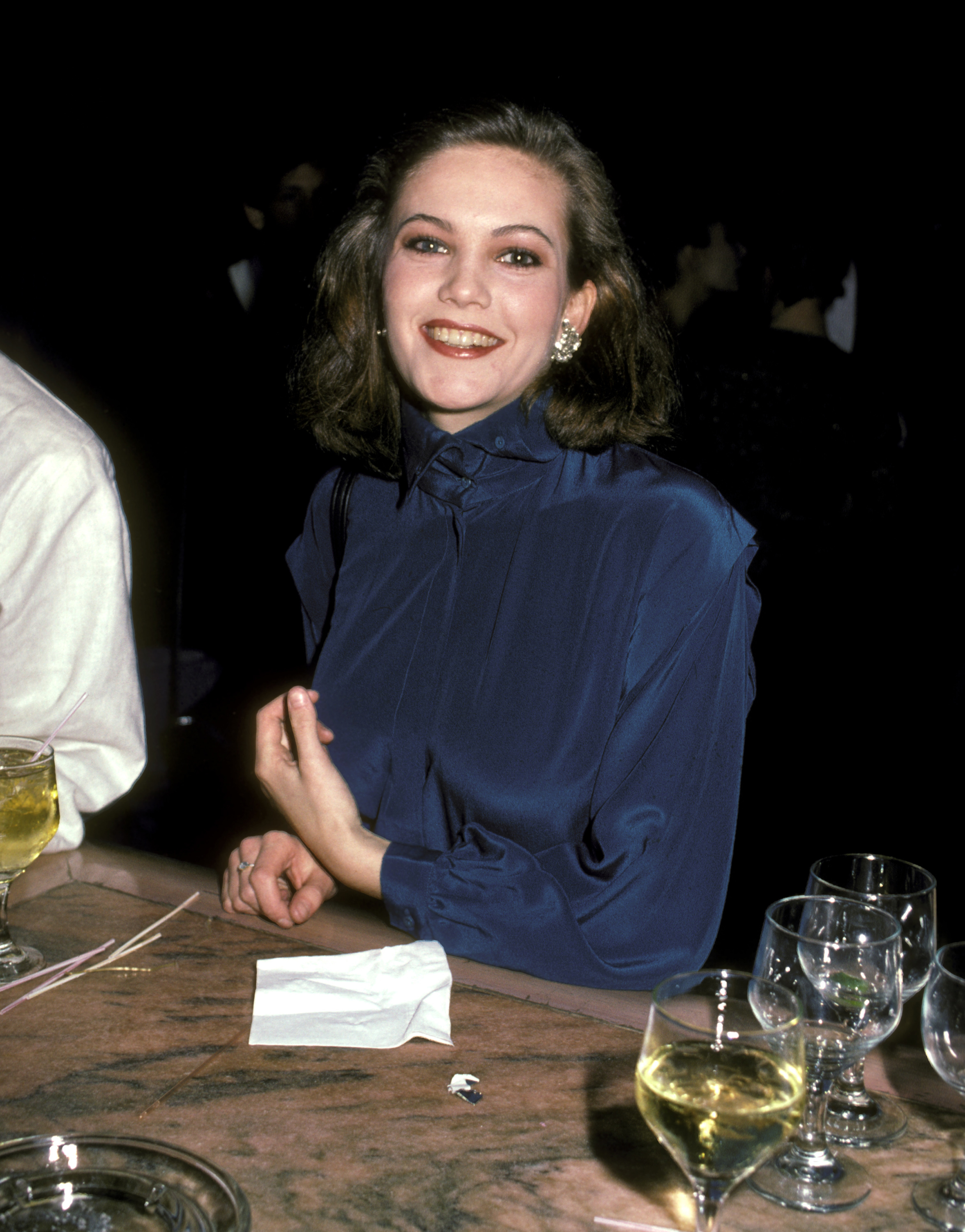 Person in elegant satin blouse smiling at an event, seated at a table with drinks in the foreground