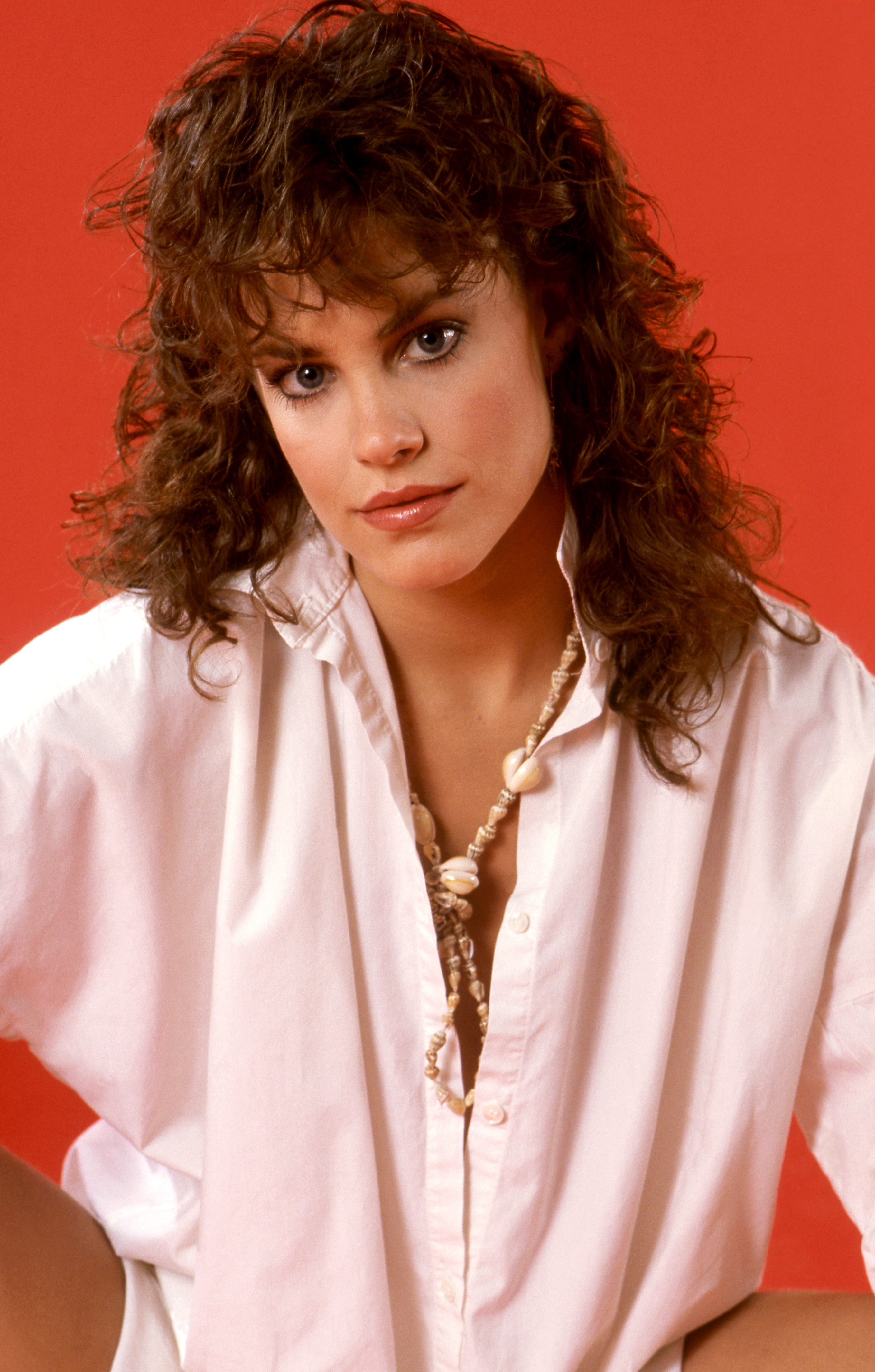 Person in 1980s fashion with curly hair and shell necklace against a plain backdrop, wearing an oversized white shirt