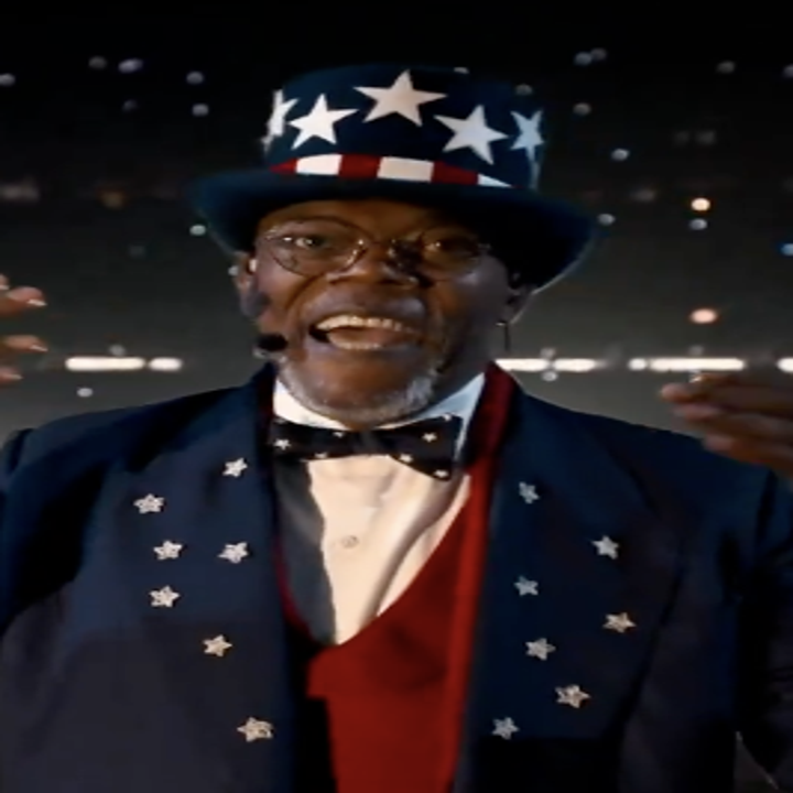 Man in a star-patterned top hat, glasses, and star-embellished suit gestures energetically on stage with a headset microphone
