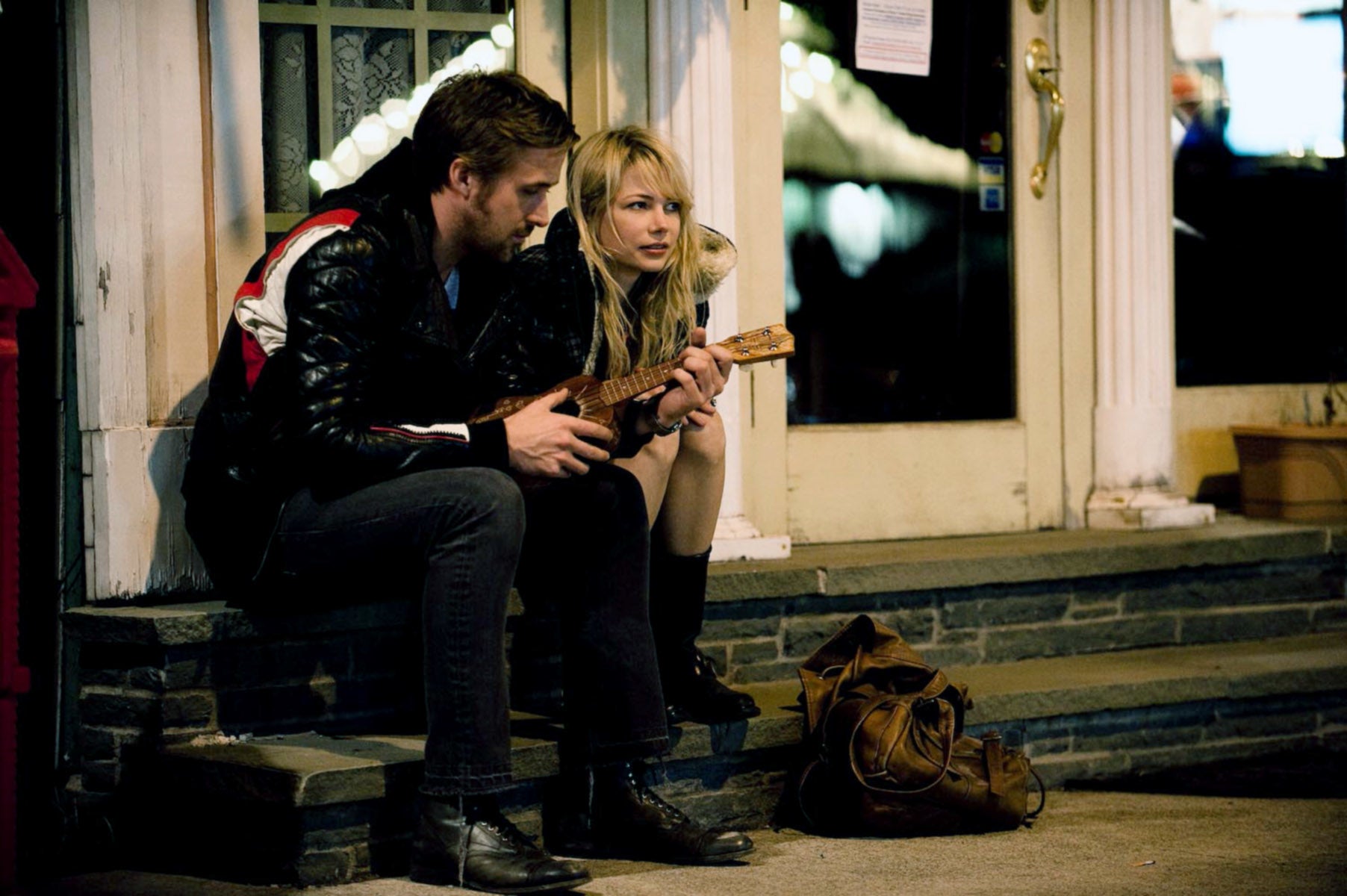 Two people sit on steps outside a building, one holding a small guitar-like instrument. Both are wearing casual clothes and leather jackets