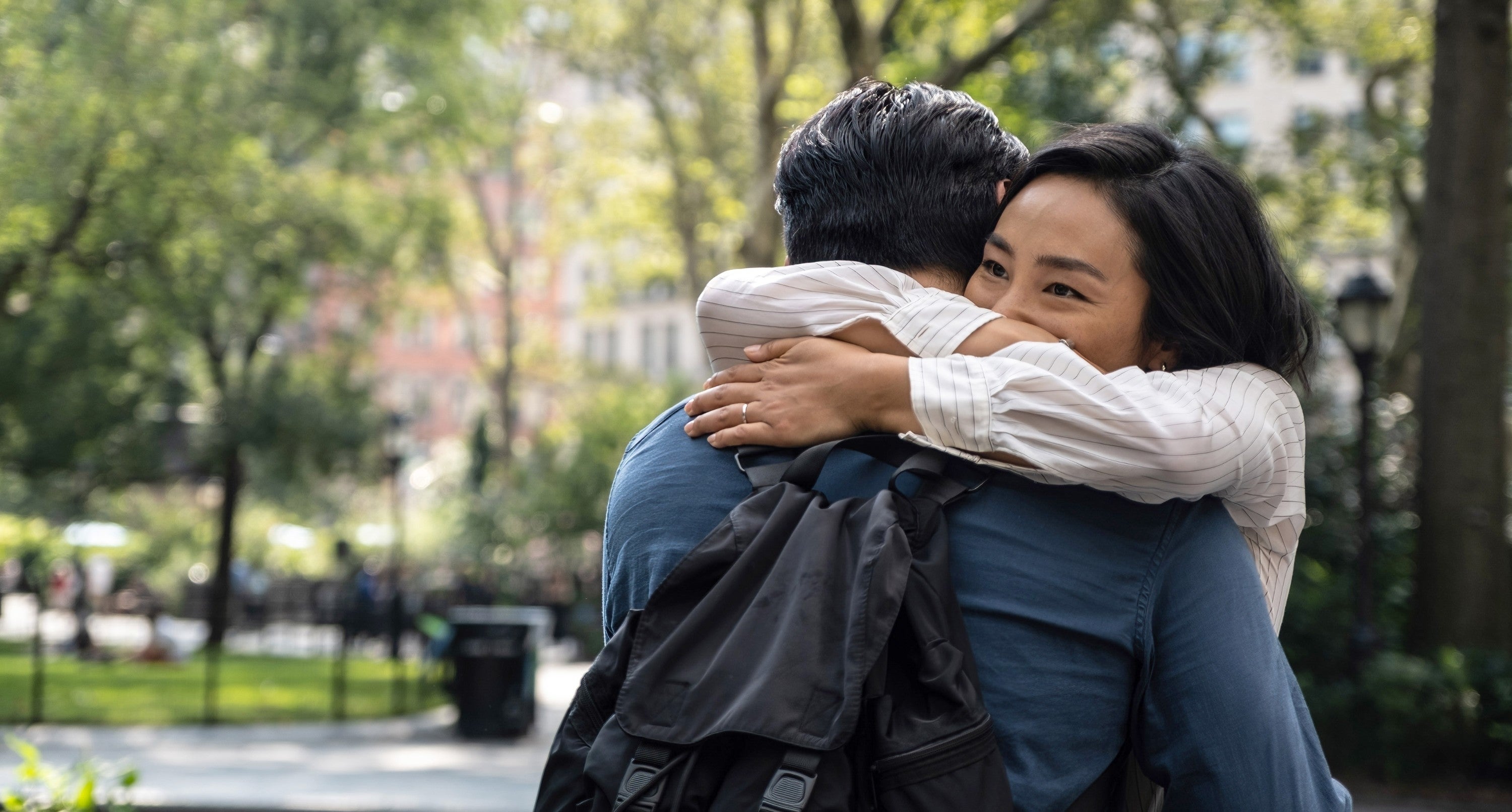 Two people warmly embrace in a park, one person smiling over the other's shoulder, conveying affection and joy in a natural setting