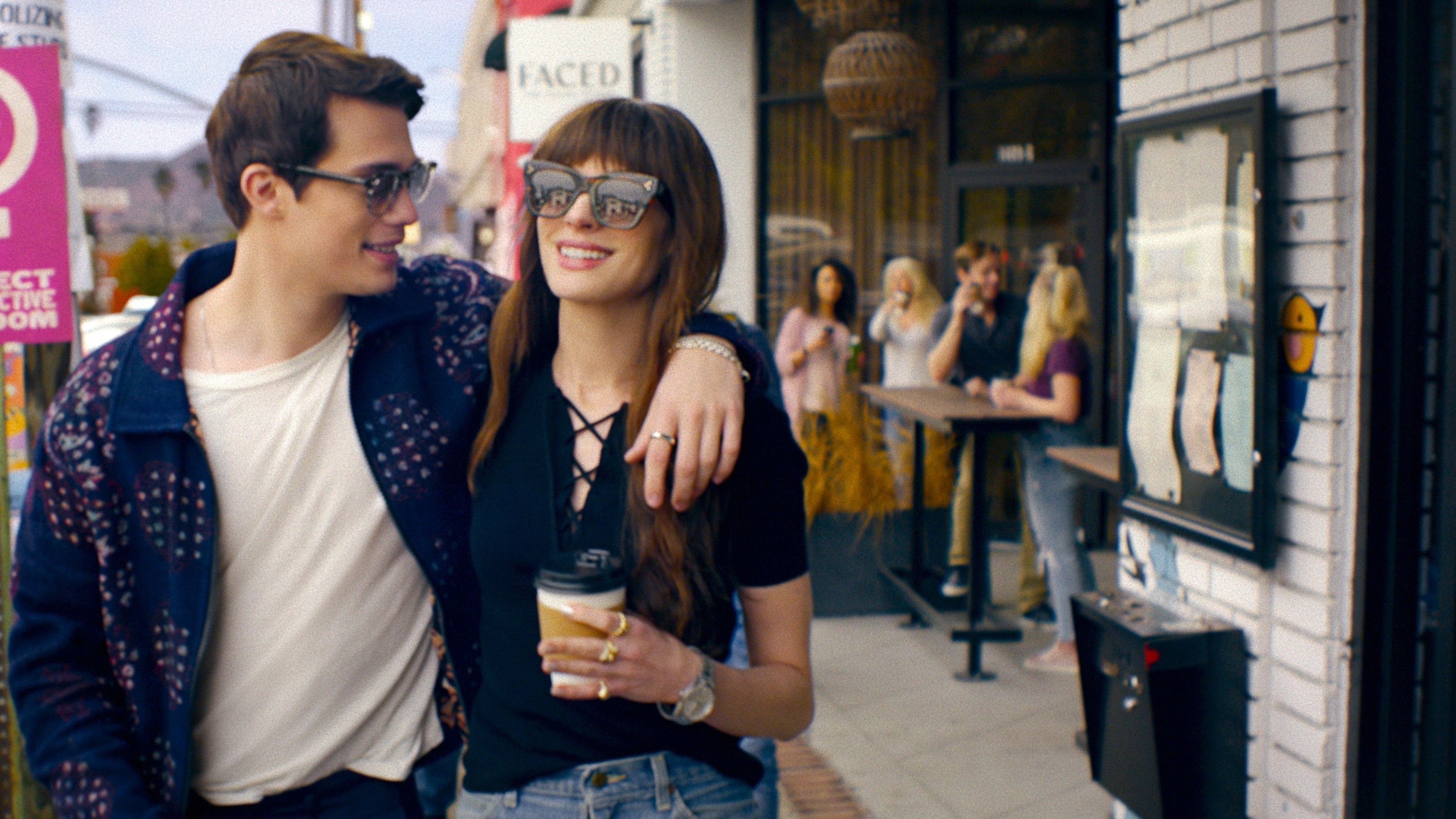 Two people walking outside a cafe, casually dressed. One is holding a coffee, both smiling and wearing sunglasses