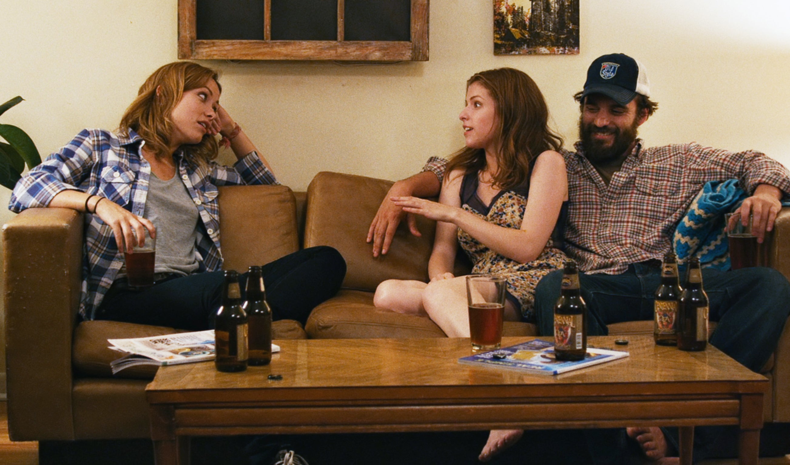 Three people relax on a couch, engaged in conversation. Beverages are on the table. Casual, homely setting with a warm atmosphere