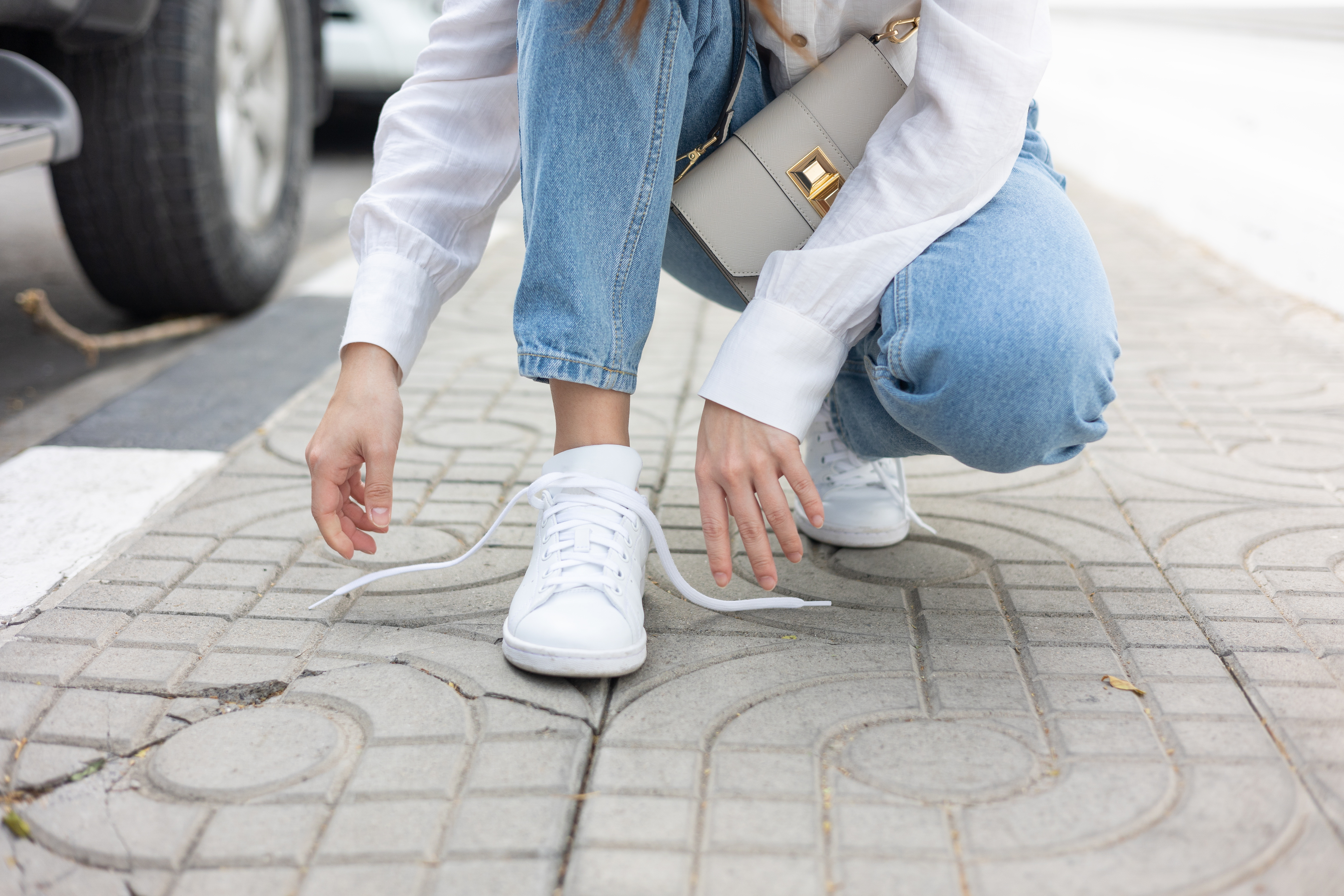 Person som huker på et fortau, binder hvite joggesko, har på seg en hvit genser og jeans, holder en lysfarget veske. Uformell og avslappet scene
